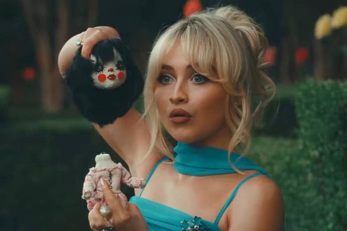 Sabrina Carpenter in a scene holding a headless doll and a doll's head with exaggerated red cheeks, wearing an elegant blue dress
