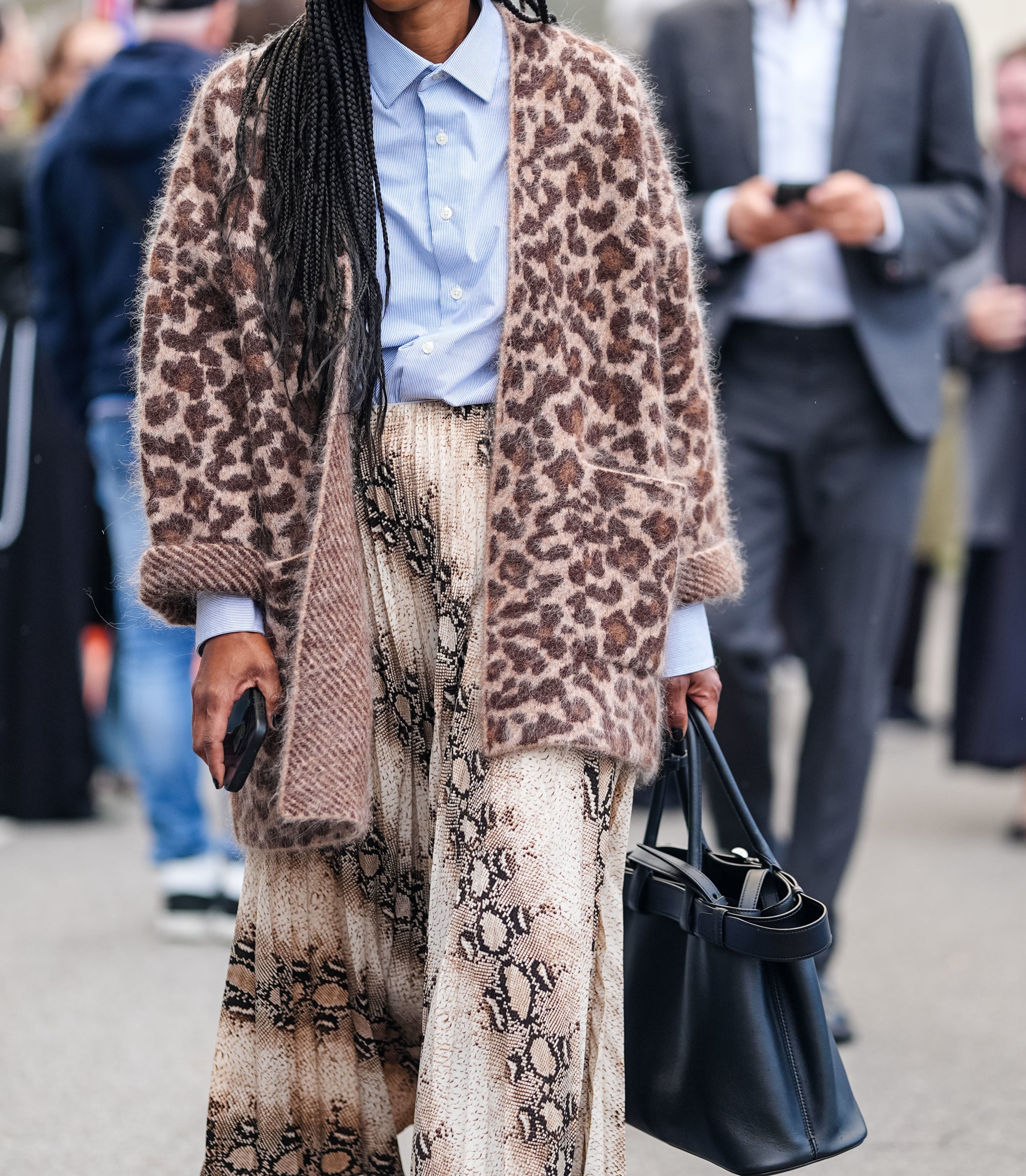 Woman in a patterned coat, blue shirt, and a snakeprint skirt walking with a black bag during a street fashion event