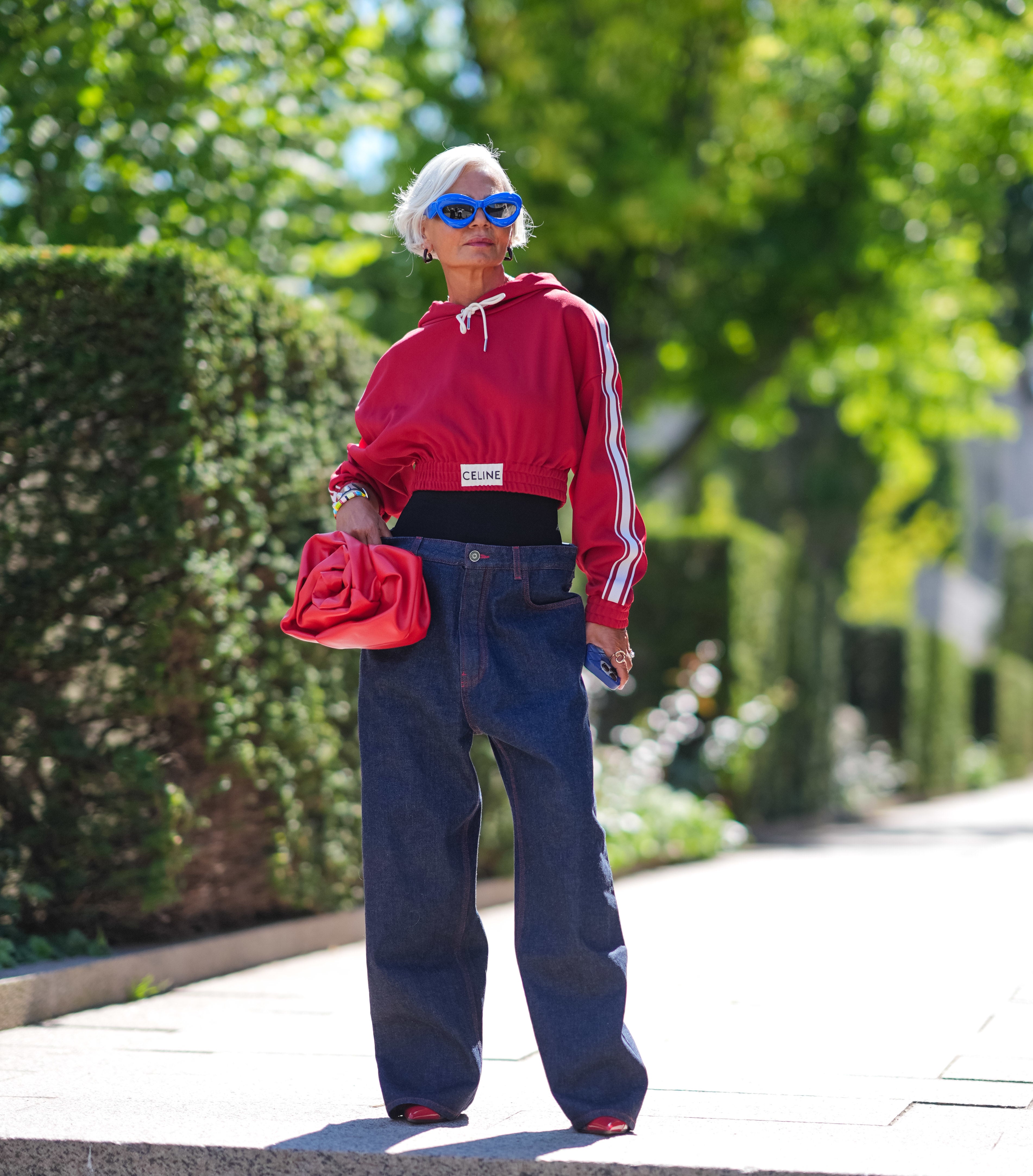 Grece Ghanem wearing a red sweater with sporty stripes, high-waisted dark jeans, and blue sunglasses, holding a red bag