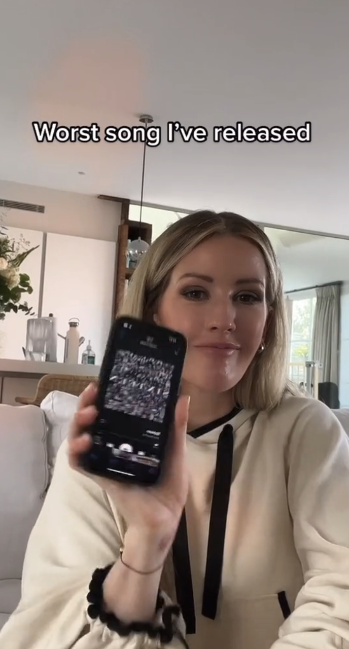 Ellie Goulding holds a smartphone with "Sixteen" artwork and smiles at the camera. Text on the image reads, "Worst song I've released."