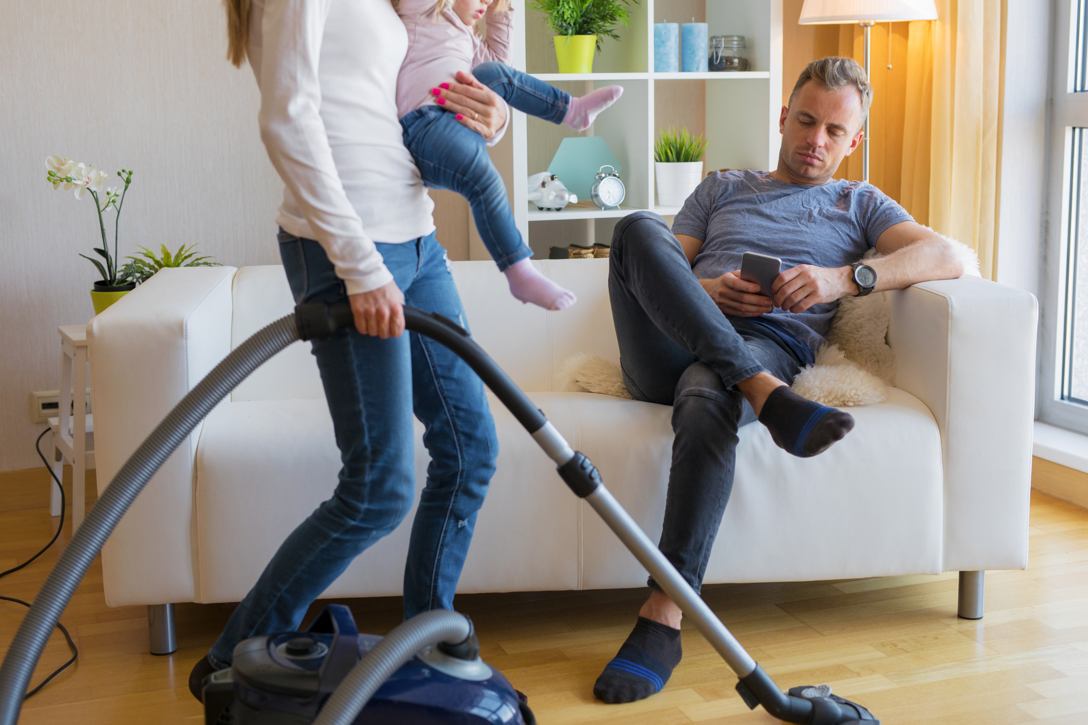 A person vacuums the living room while holding a child. Another person sits on a couch, looking at a smartphone