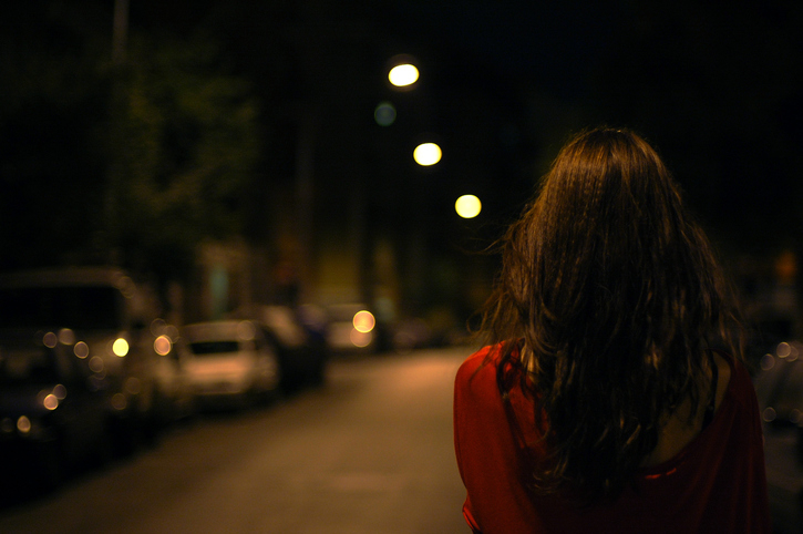 Person with long hair walks alone on a dimly lit street at night, wearing a red top. The street is lined with parked cars