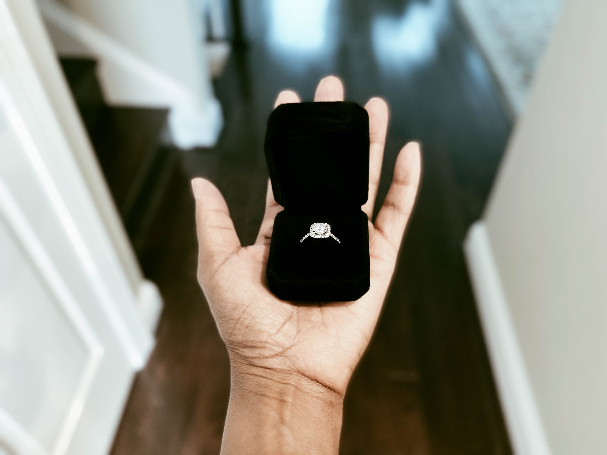 A person holds a black velvet ring box containing a diamond engagement ring, showing it off in a hallway
