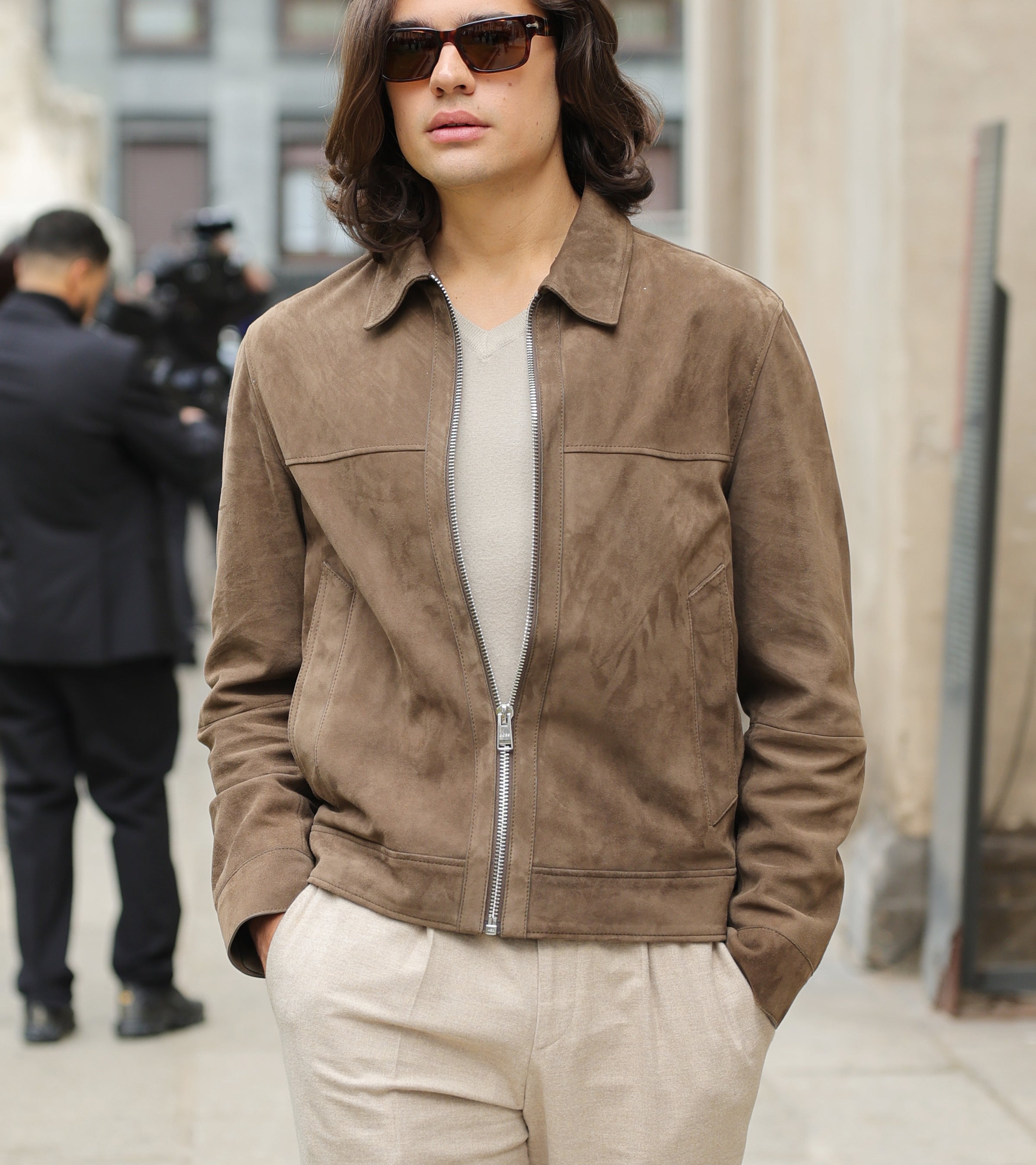 Person in a suede zip-up jacket, sunglasses, and beige pants, walking on a street