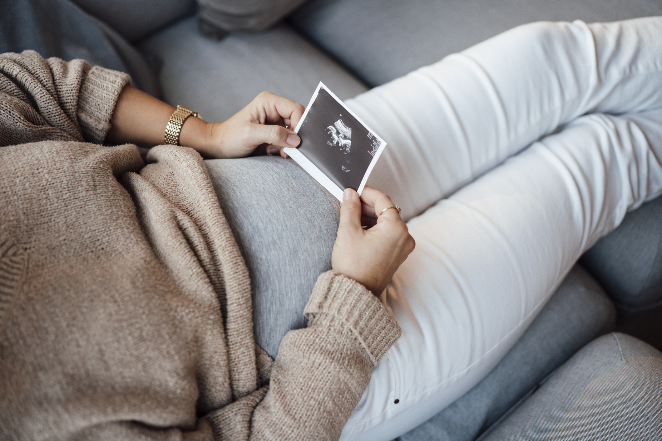A person rests on a couch holding a sonogram image over their stomach, suggesting pregnancy. They are wearing a beige sweater and white pants