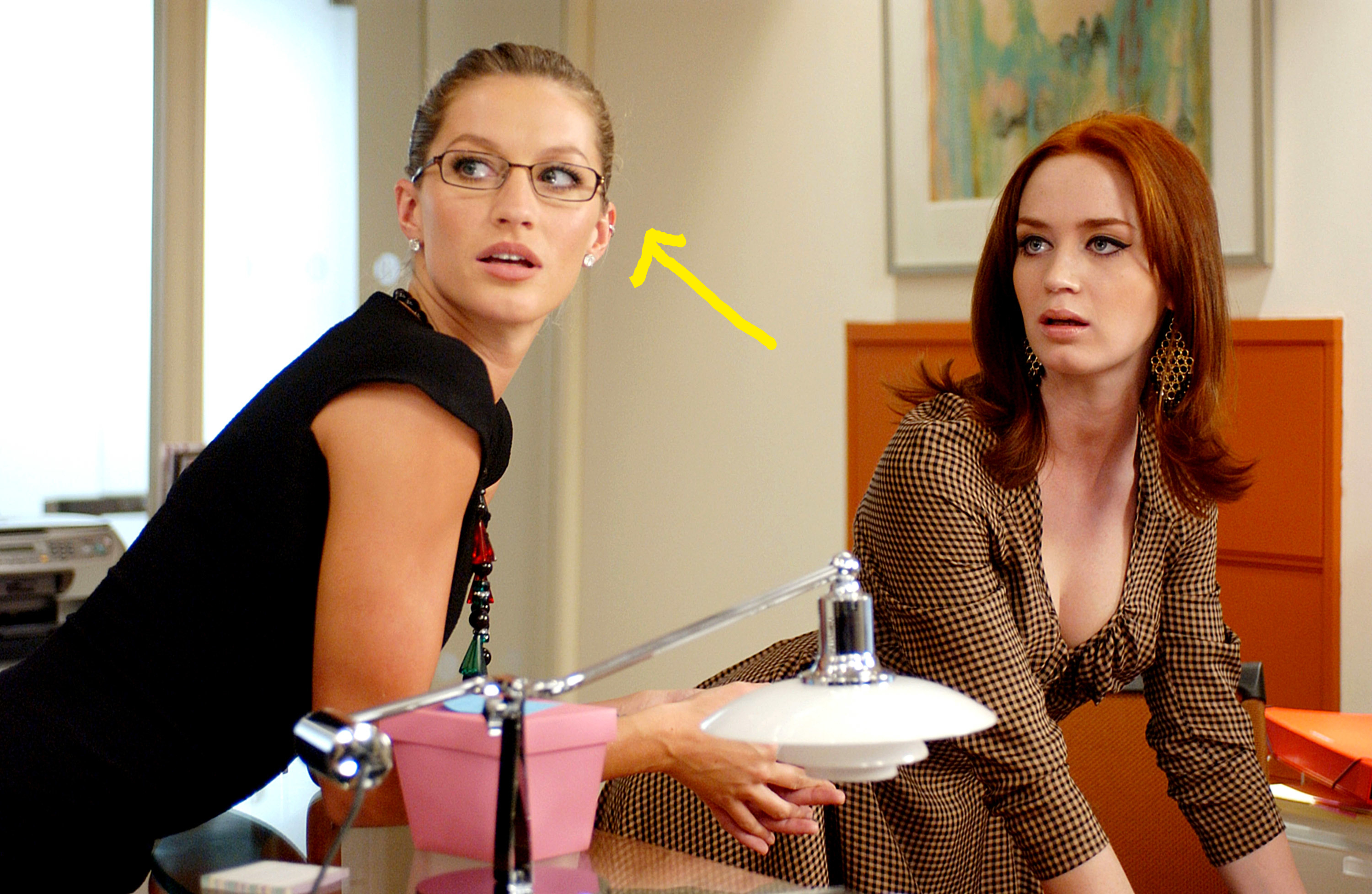 Gisele Bündchen and Emily Blunt in a scene from "The Devil Wears Prada," both looking off to the side, standing at a desk with office supplies in the foreground