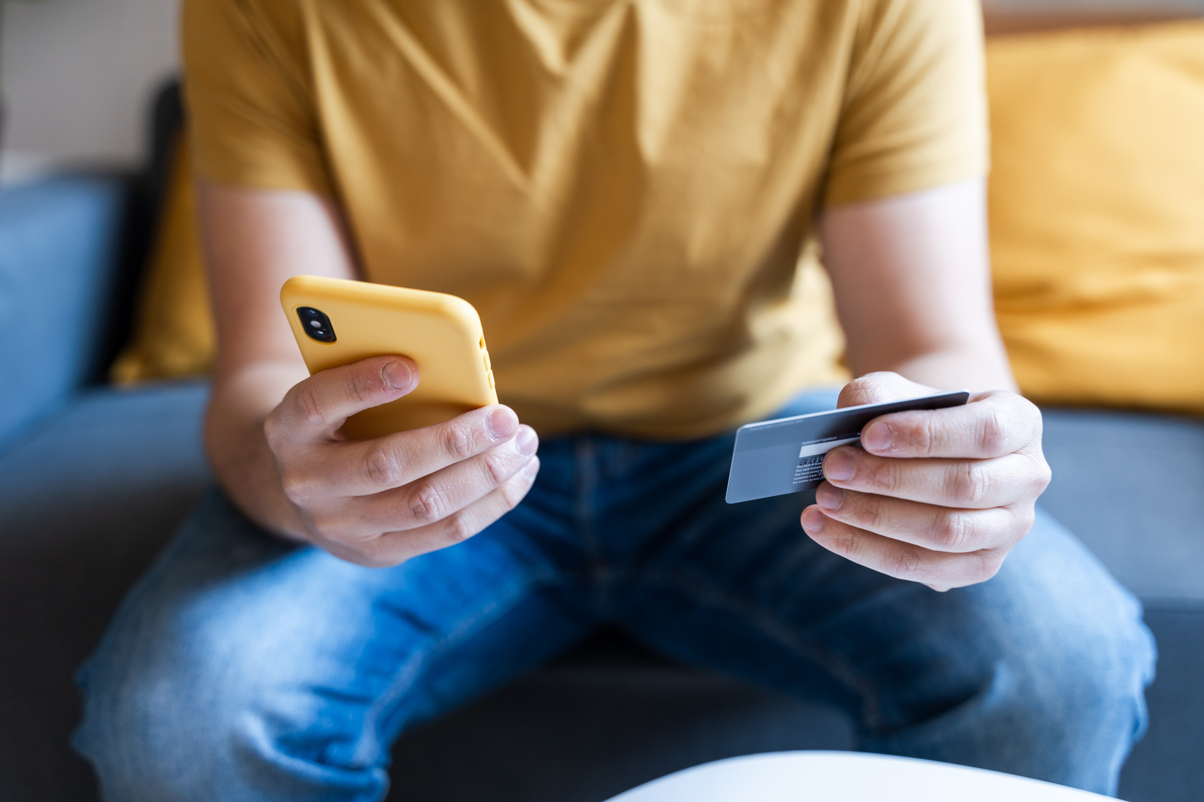 A person wearing a yellow shirt is sitting on a couch, holding a smartphone in one hand and a credit card in the other, likely making an online purchase
