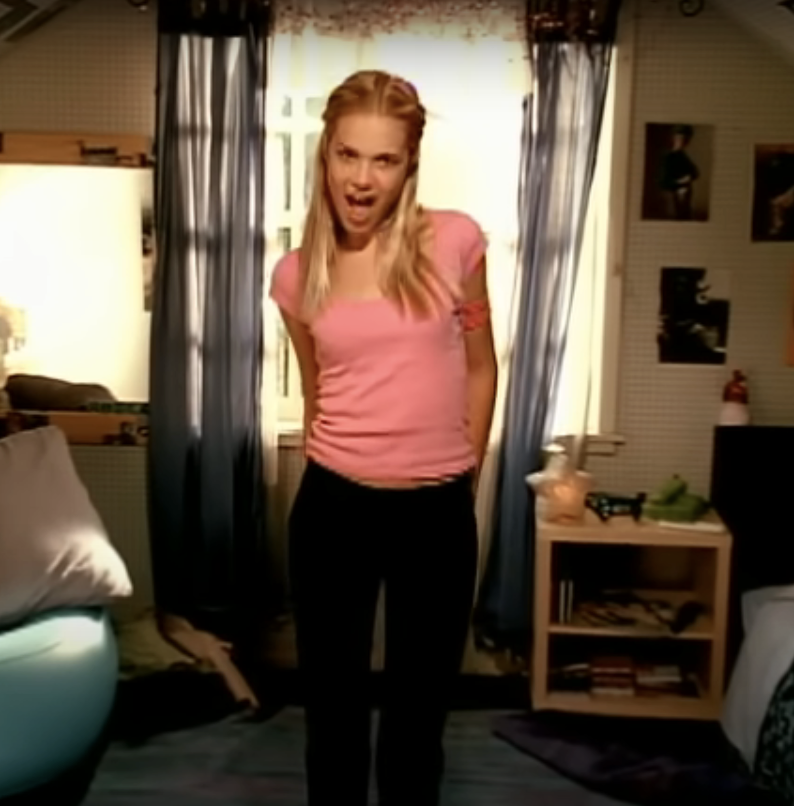 Mandy Moore stands in a casual indoor setting, wearing a short-sleeved shirt and pants, with her hands behind her back, smiling at the camera