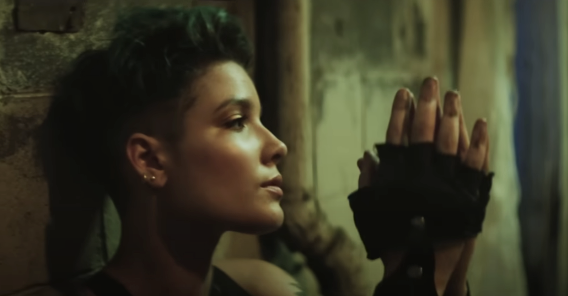 Halsey wearing fingerless gloves, leaning against a wall, and clasping her hands together while looking thoughtfully ahead