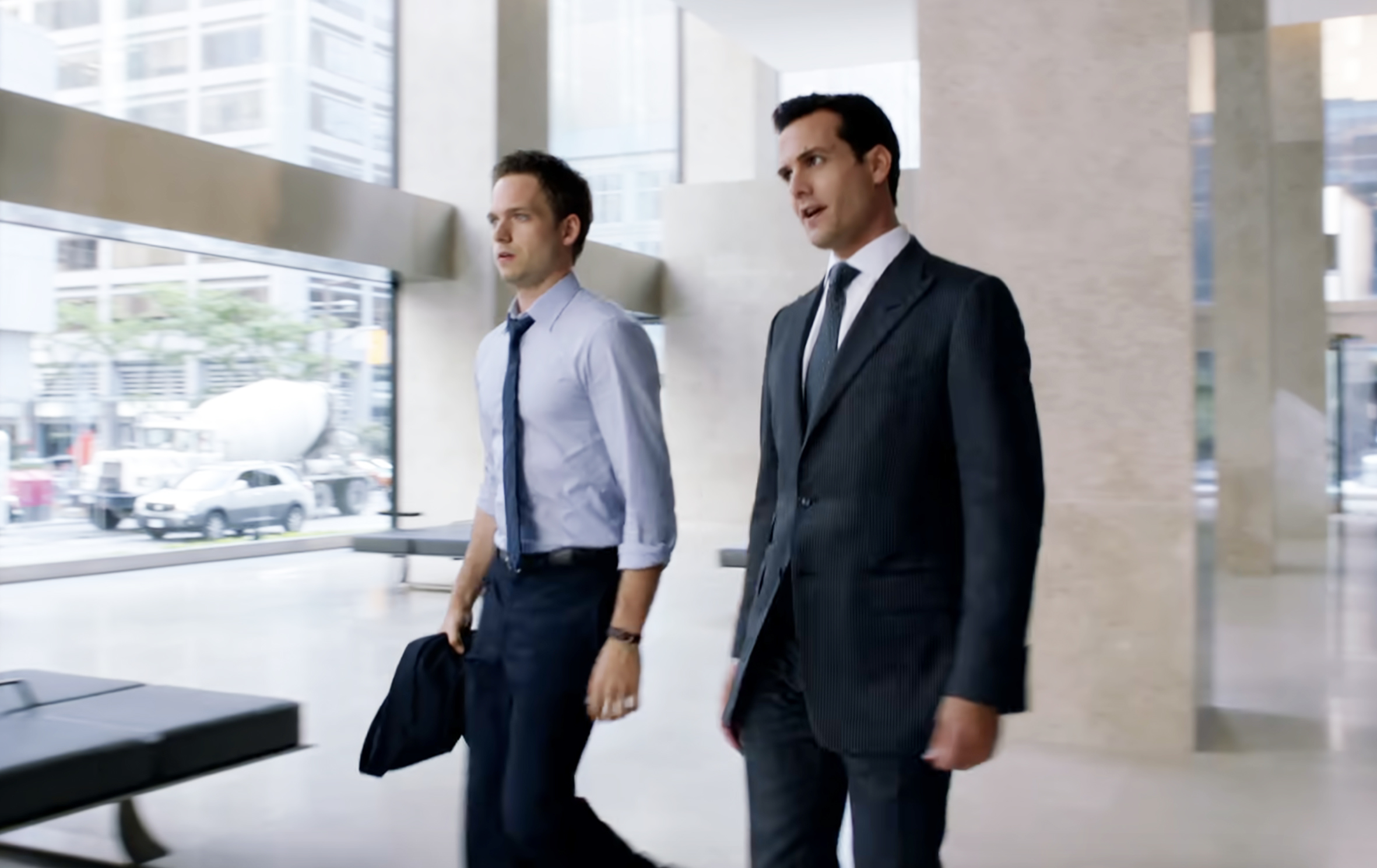 Patrick J. Adams and Gabriel Macht in a scene from the TV show "Suits," walking inside a glass-paneled office building, both wearing business attire