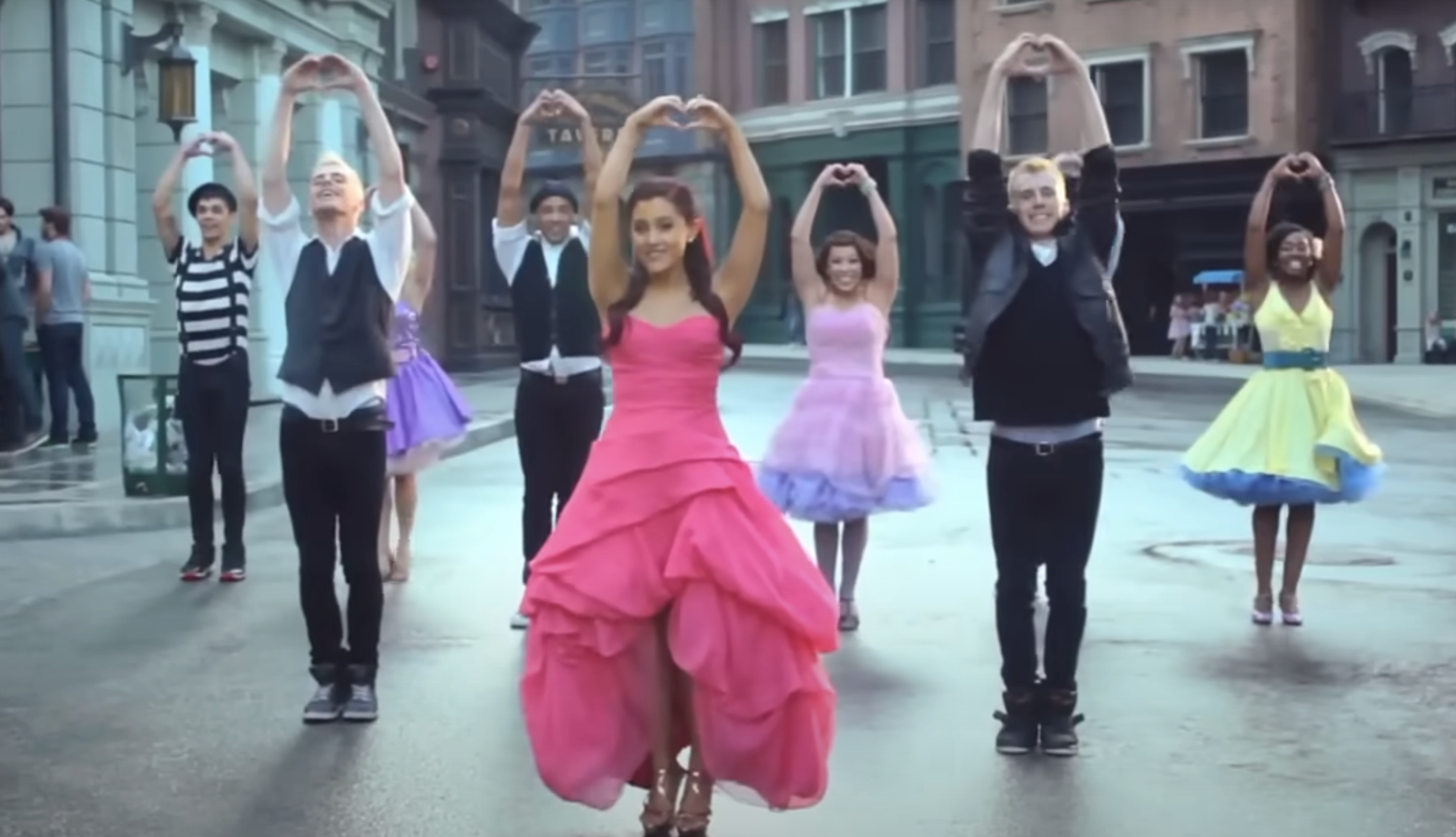 Ariana Grande and dancers pose with arms making heart shapes in a street scene, dressed in formal attire. Ariana wears a strapless, layered dress