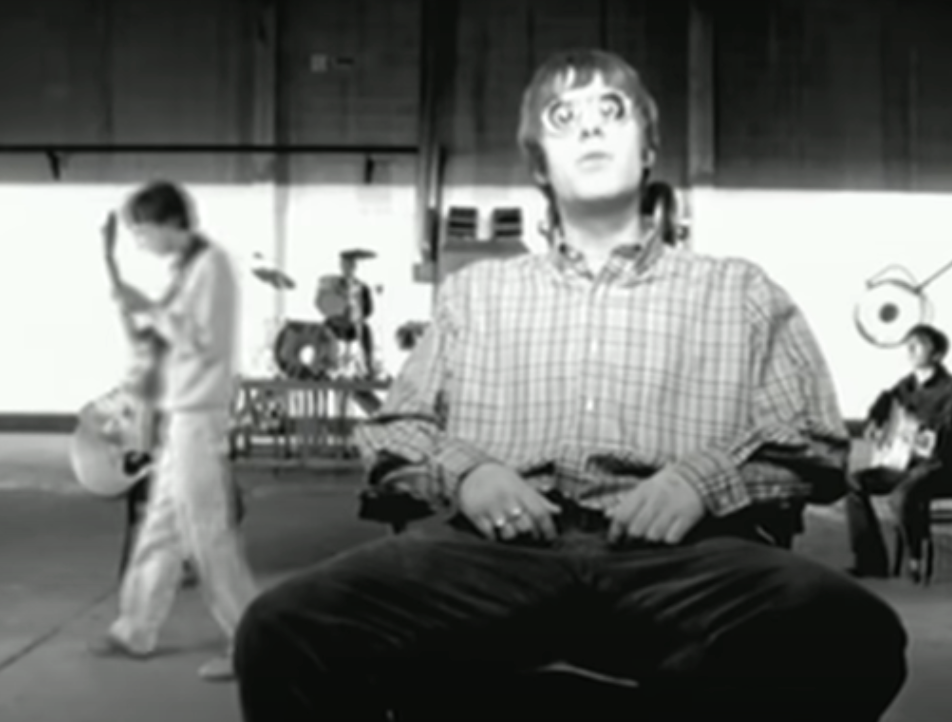 Liam Gallagher sits in a room with musical instruments in the background, appearing thoughtful, in a scene from an Oasis music video
