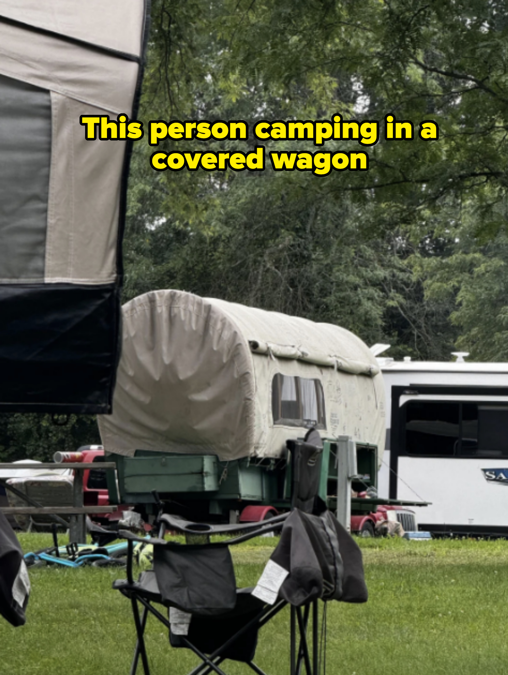 Covered wagon in a campground with chairs and tents, surrounded by trees and an RV in the background
