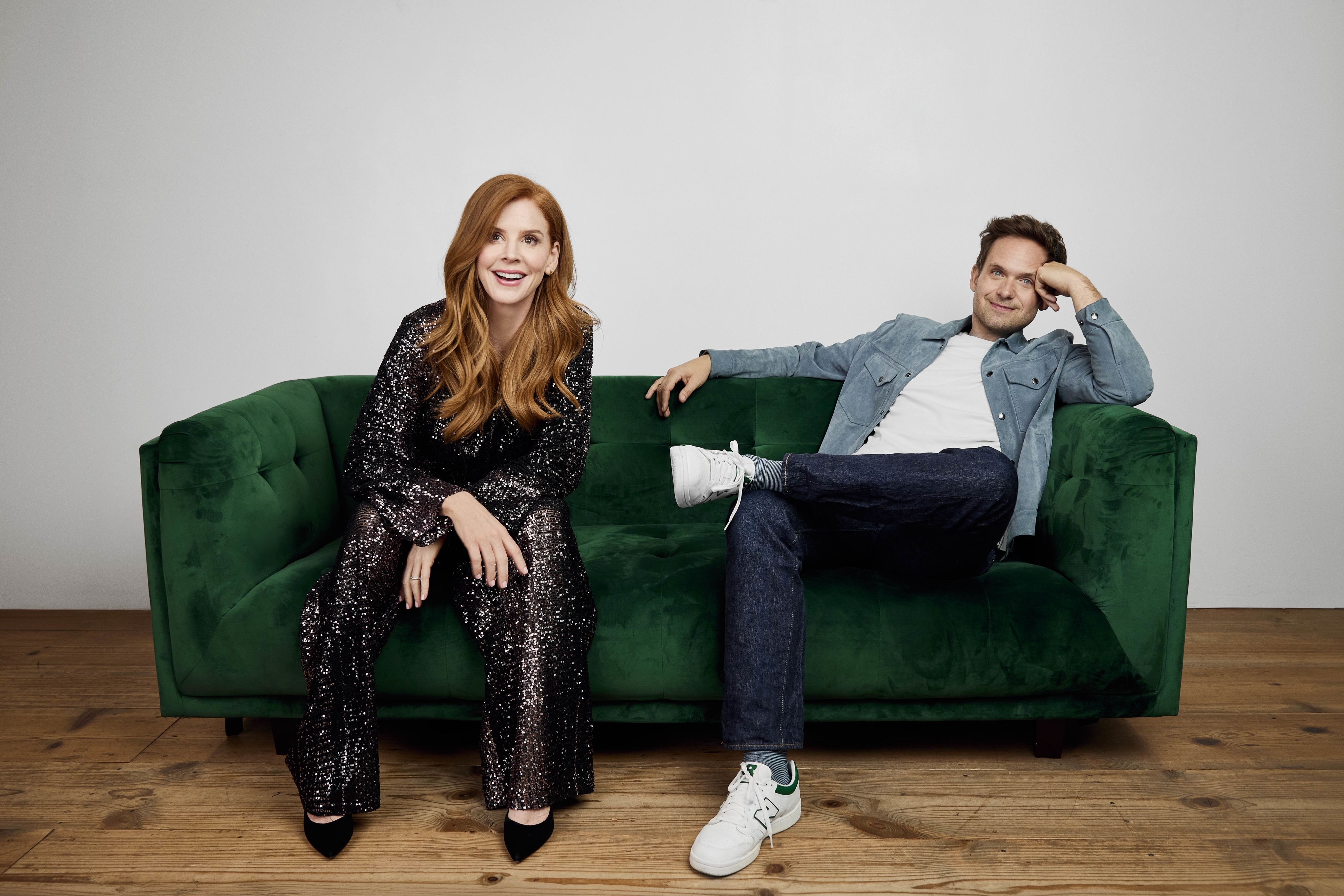 Sarah Rafferty in a sparkling jumpsuit and Patrick J. Adams in a denim jacket, jeans, and sneakers, posing on a green sofa for a photoshoot