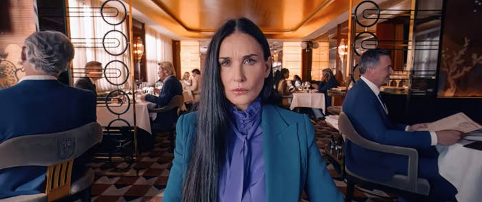 Demi Moore sits at a dining table in an upscale restaurant. She is wearing a fitted blue blazer over a high-collared purple blouse. Other diners can be seen behind her in a scene from The Substance