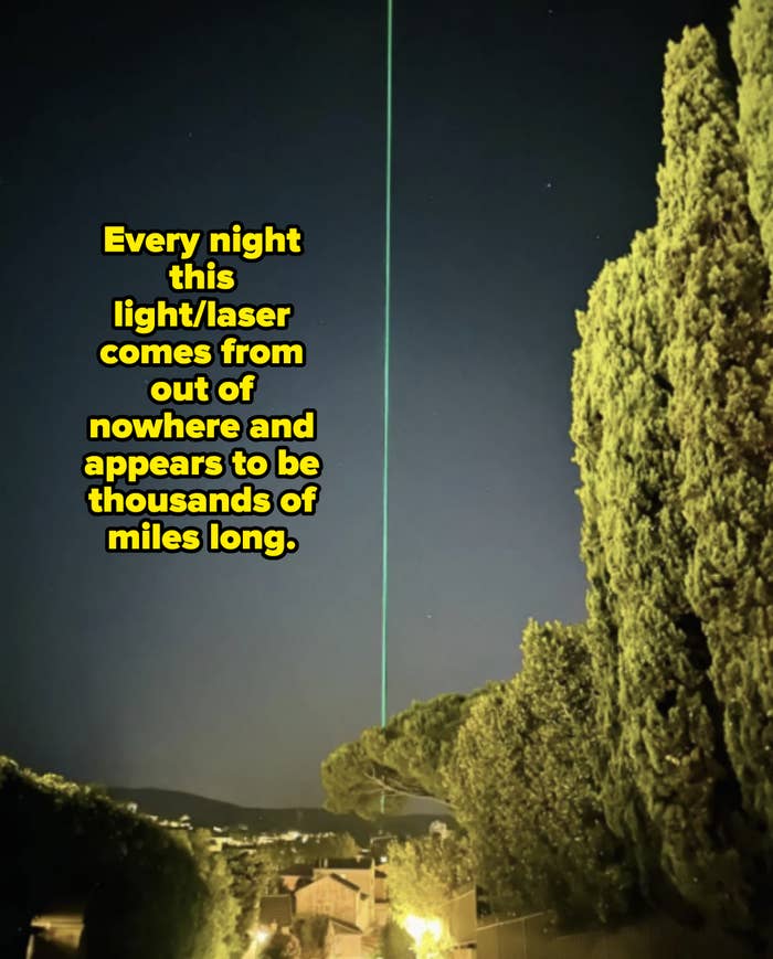 A green laser beam shines vertically into the night sky over a residential area with trees and houses below