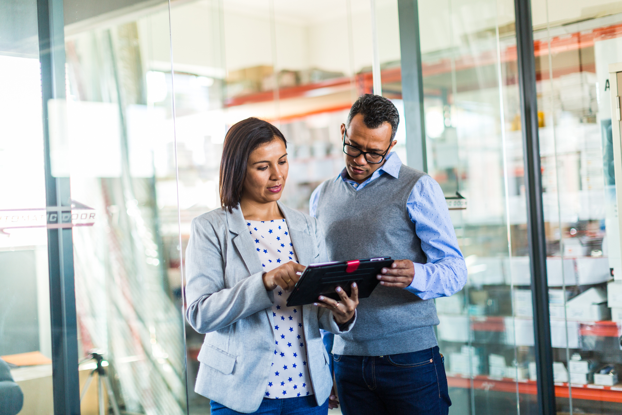 Two professionals review information on a tablet while standing in an office with glass walls and shelves in the background