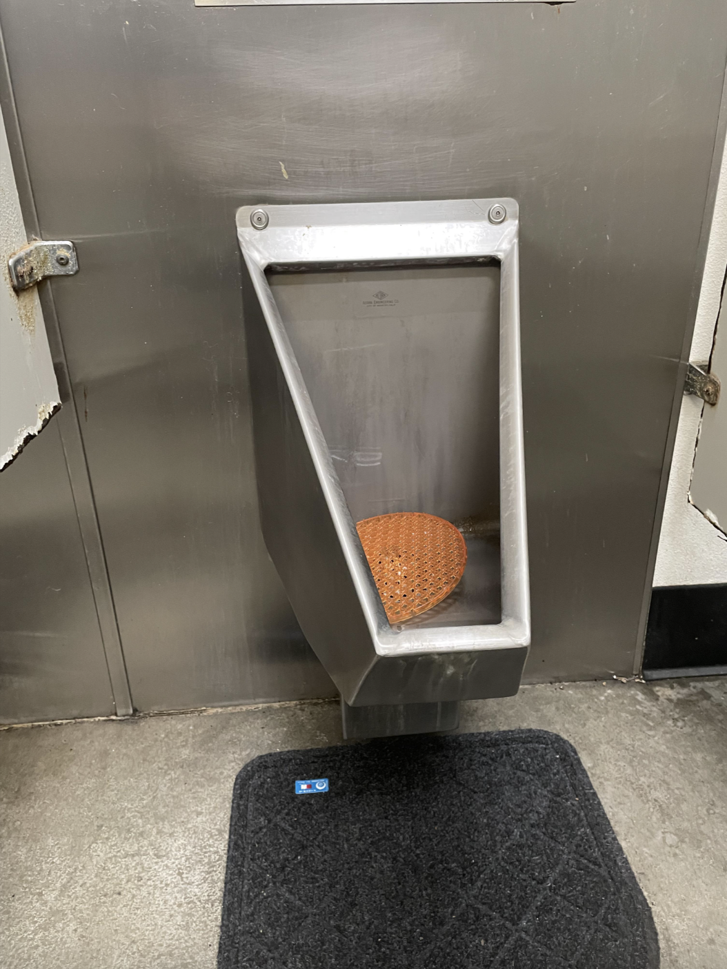 An unusual triangular urinal with a perforated copper drain mat in a public restroom, featuring an industrial design