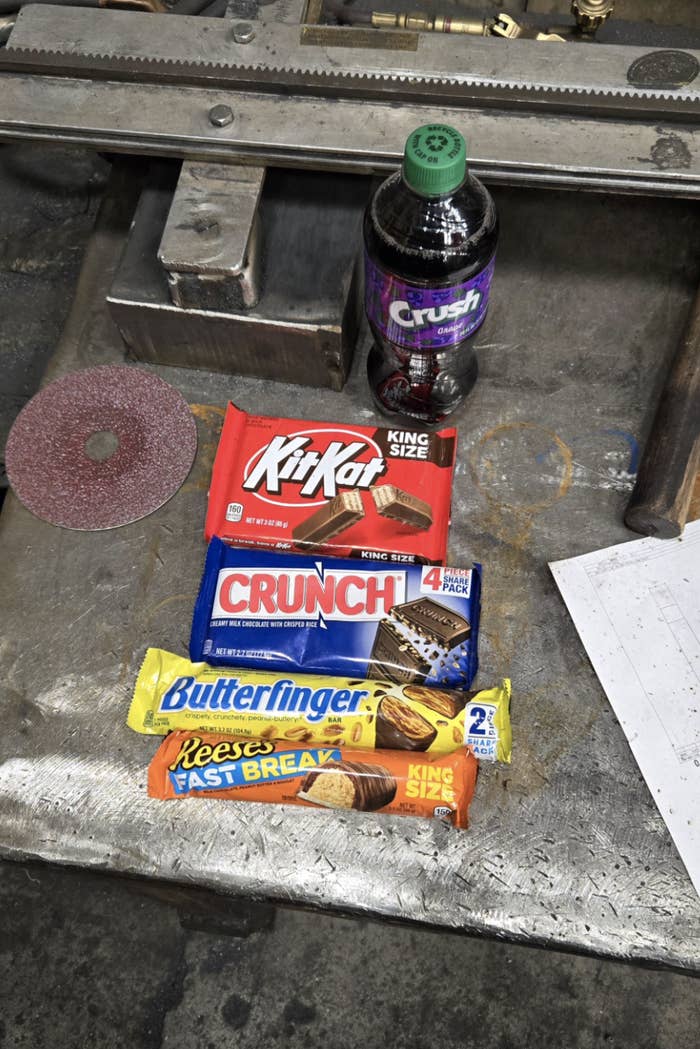 Various candy bars and a soda bottle are placed on a metal surface. The candy bars include Kit Kat, Crunch, Butterfinger, and Reese's Fast Break. The soda is a Grape Crush