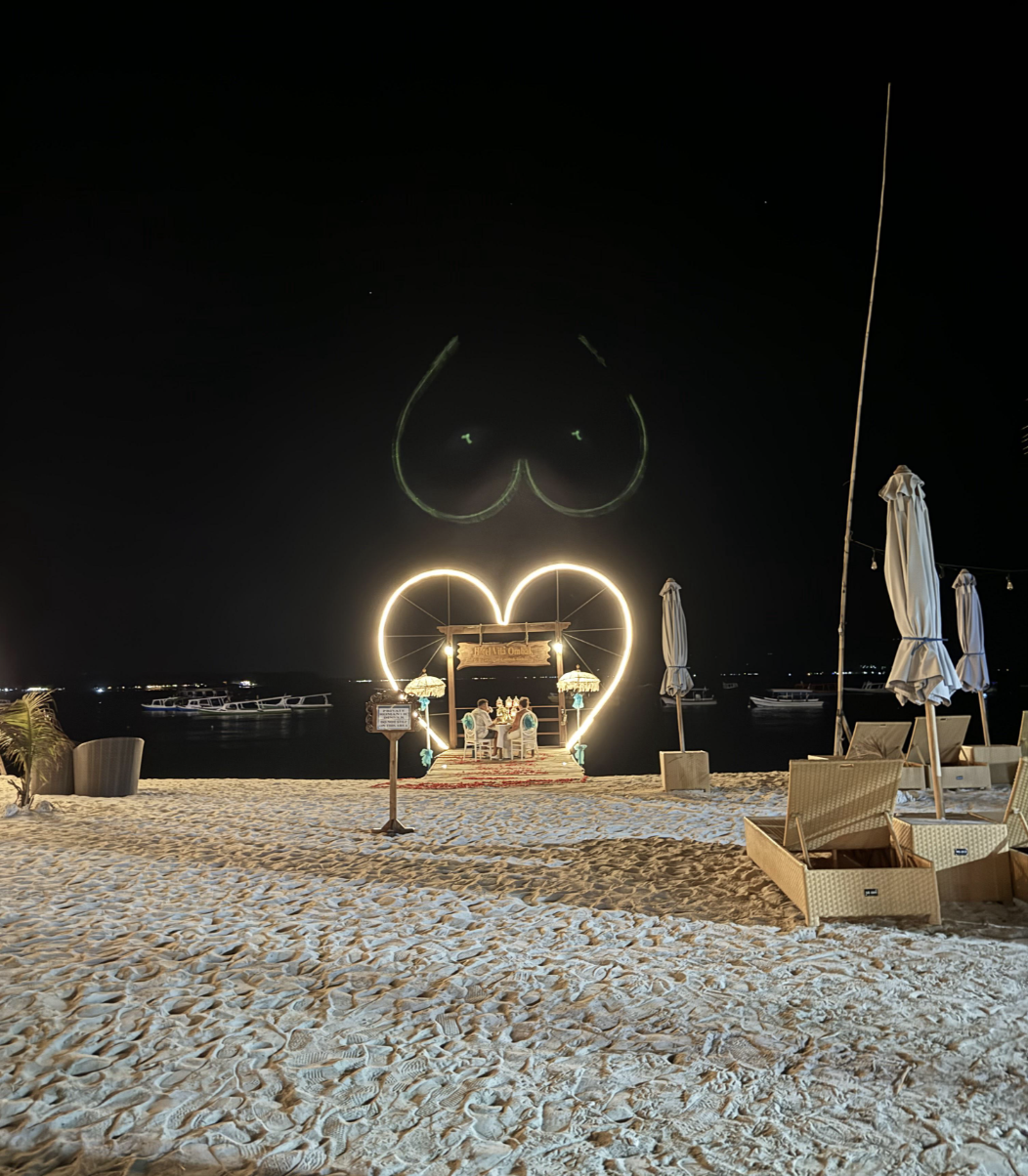 Beachfront bar decorated with heart-shaped lighting, under a dark sky with boats in the background. No people are visible in the image
