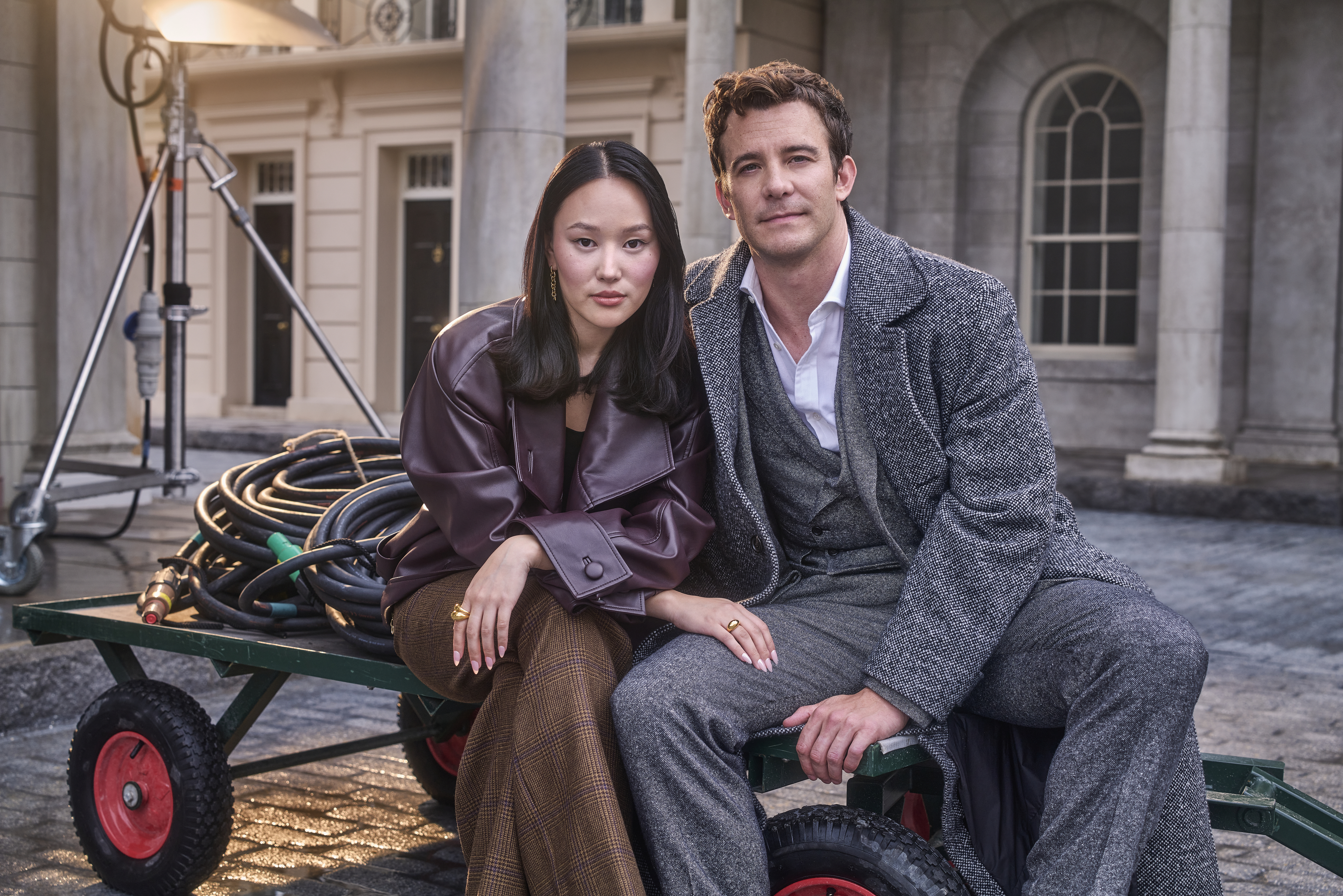 Simone Ashley and Jonathan Bailey sit side-by-side on a set, dressed in stylish, period-inspired outfits