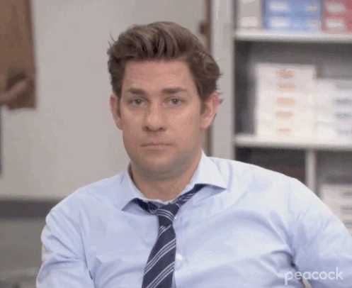 John Krasinski, dressed in a blue shirt and striped tie, sitting in an office setting, staring at the camera with a neutral expression