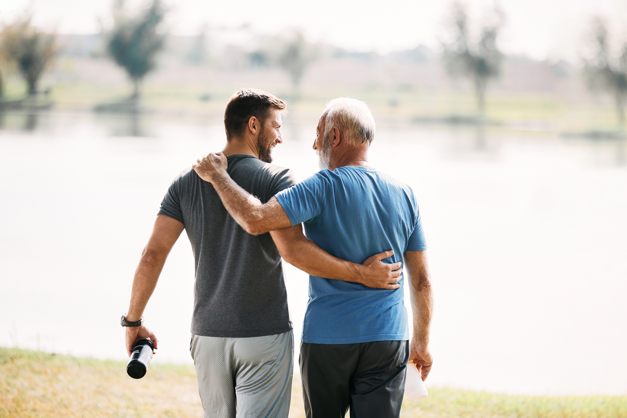 Two men, one younger holding a water bottle and another older, walk by a lakeside with arms around each other, smiling and sharing a moment