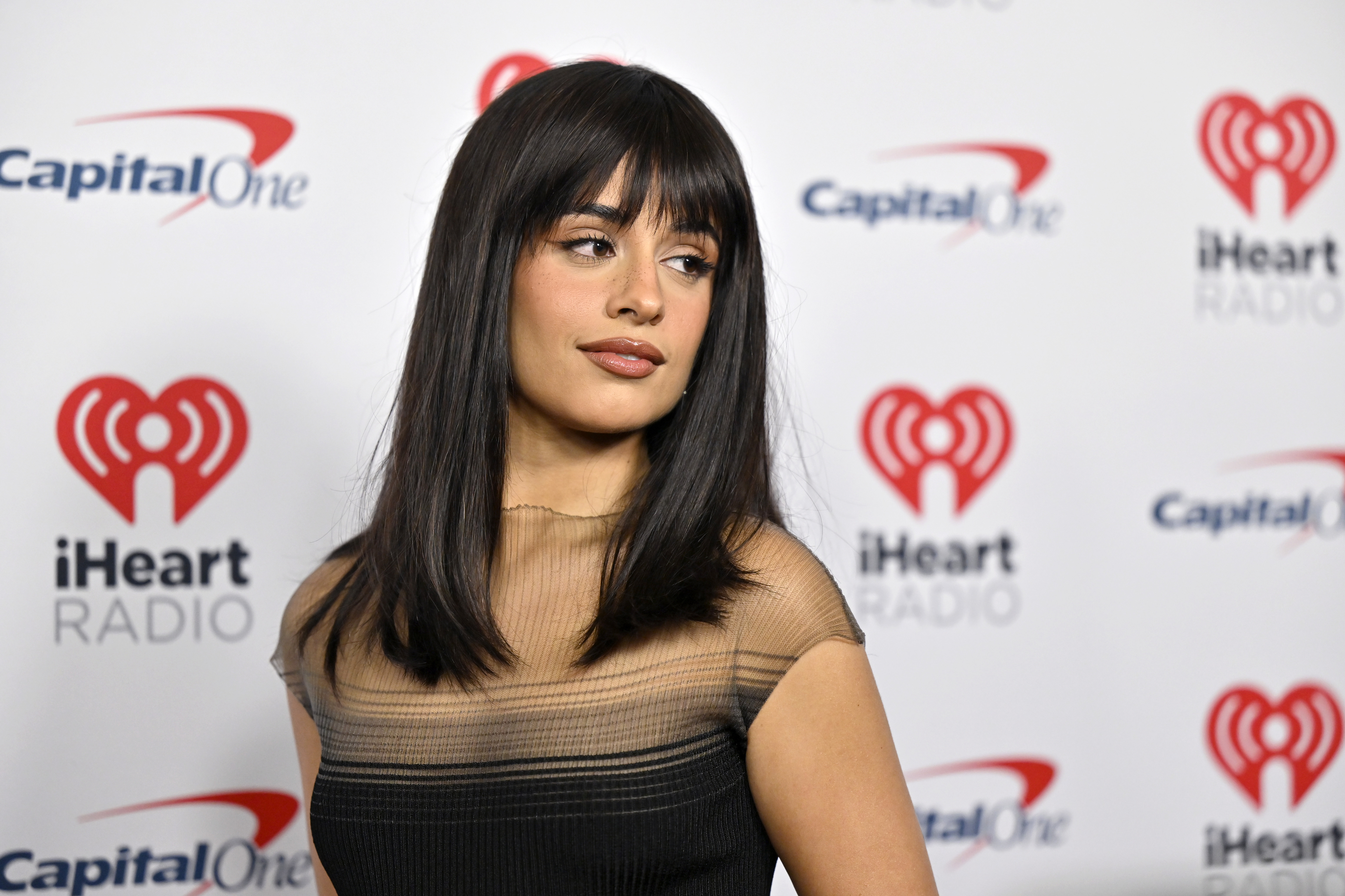 Camila Cabello poses in a sheer, short-sleeved dress at an iHeartRadio event with Capital One logos in the background