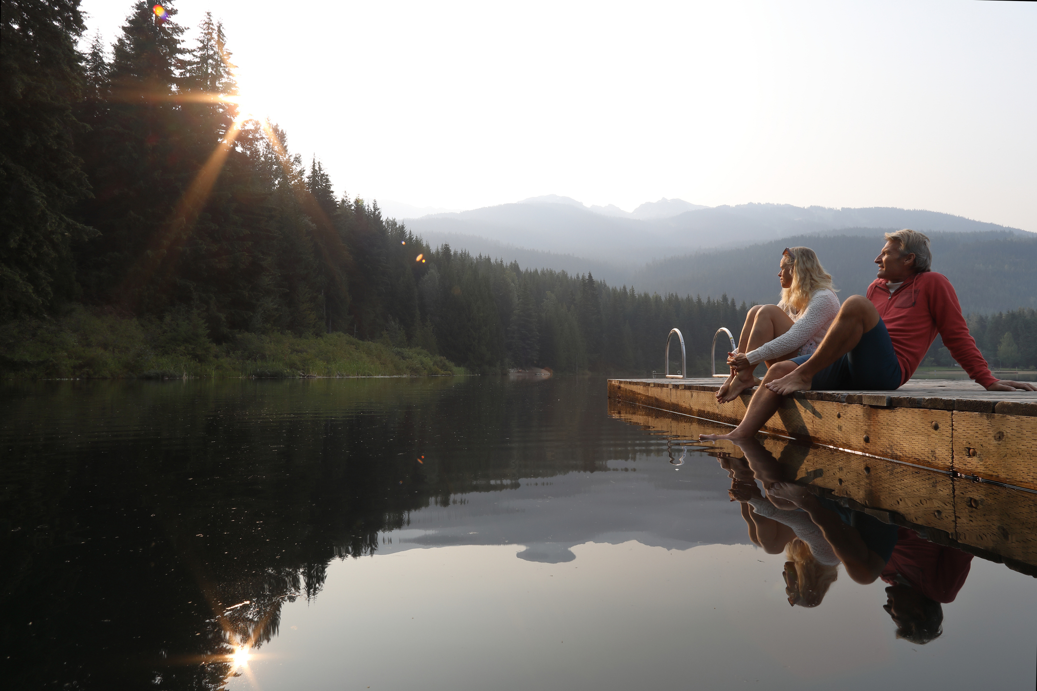 A woman and man sit at the edge of a wooden dock overlooking a serene lake surrounded by mountains and trees during sunrise or sunset