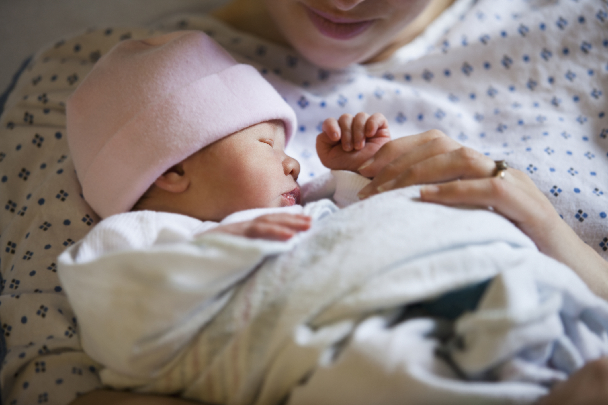 A newborn baby with a pink hat sleeps cradled in a woman's arms, who is wearing a hospital gown