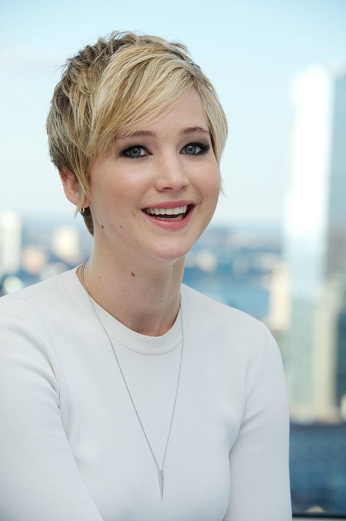Jennifer Lawrence smiles while wearing a simple, elegant top and a delicate necklace. She has a blonde pixie cut