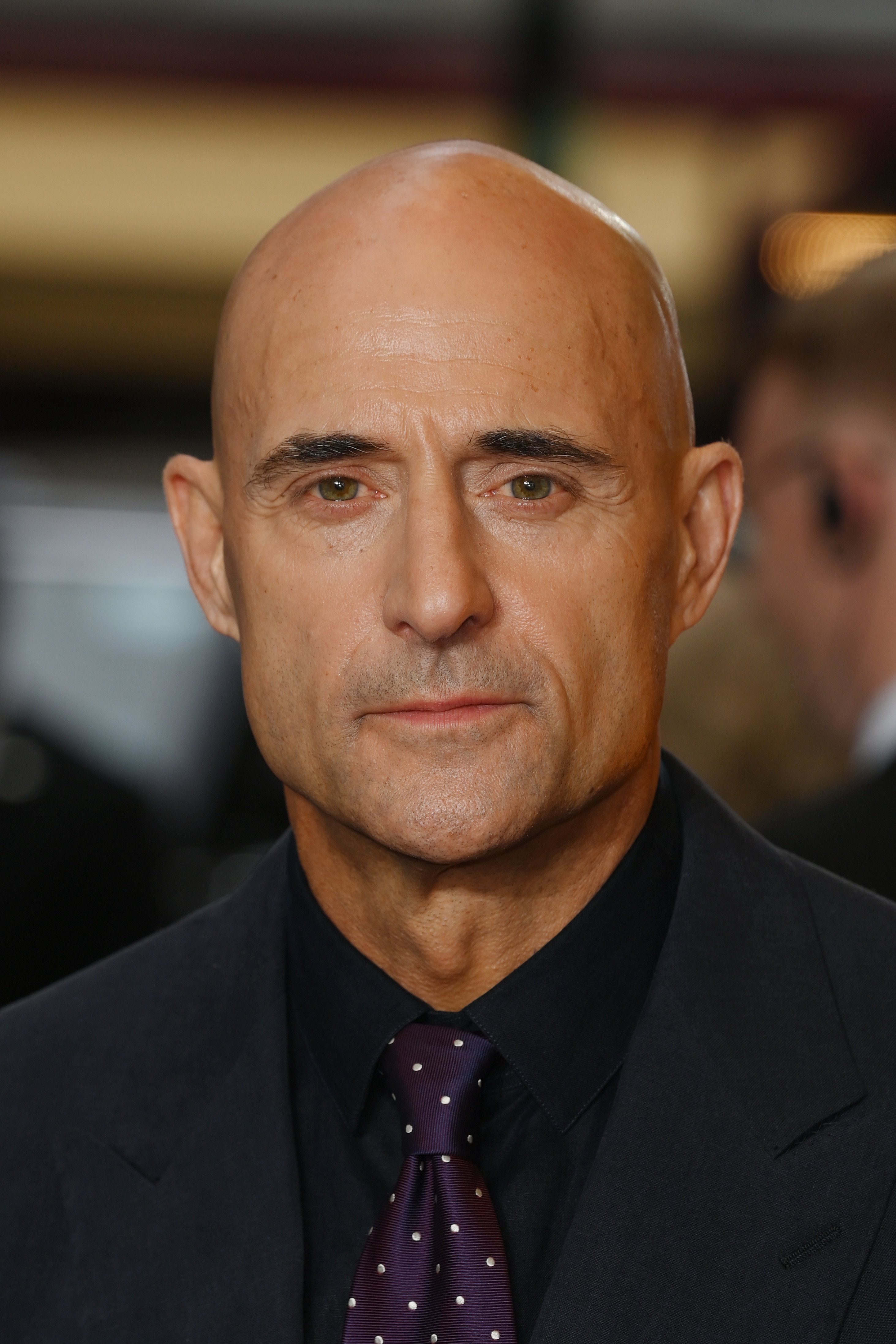 Mark Strong wears a dark suit and a dotted tie, posing at an event