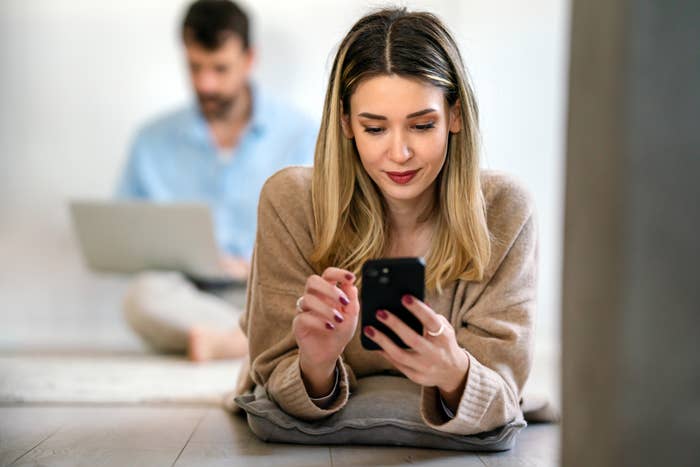 A woman lies on the floor using a smartphone while a man in the background works on a laptop