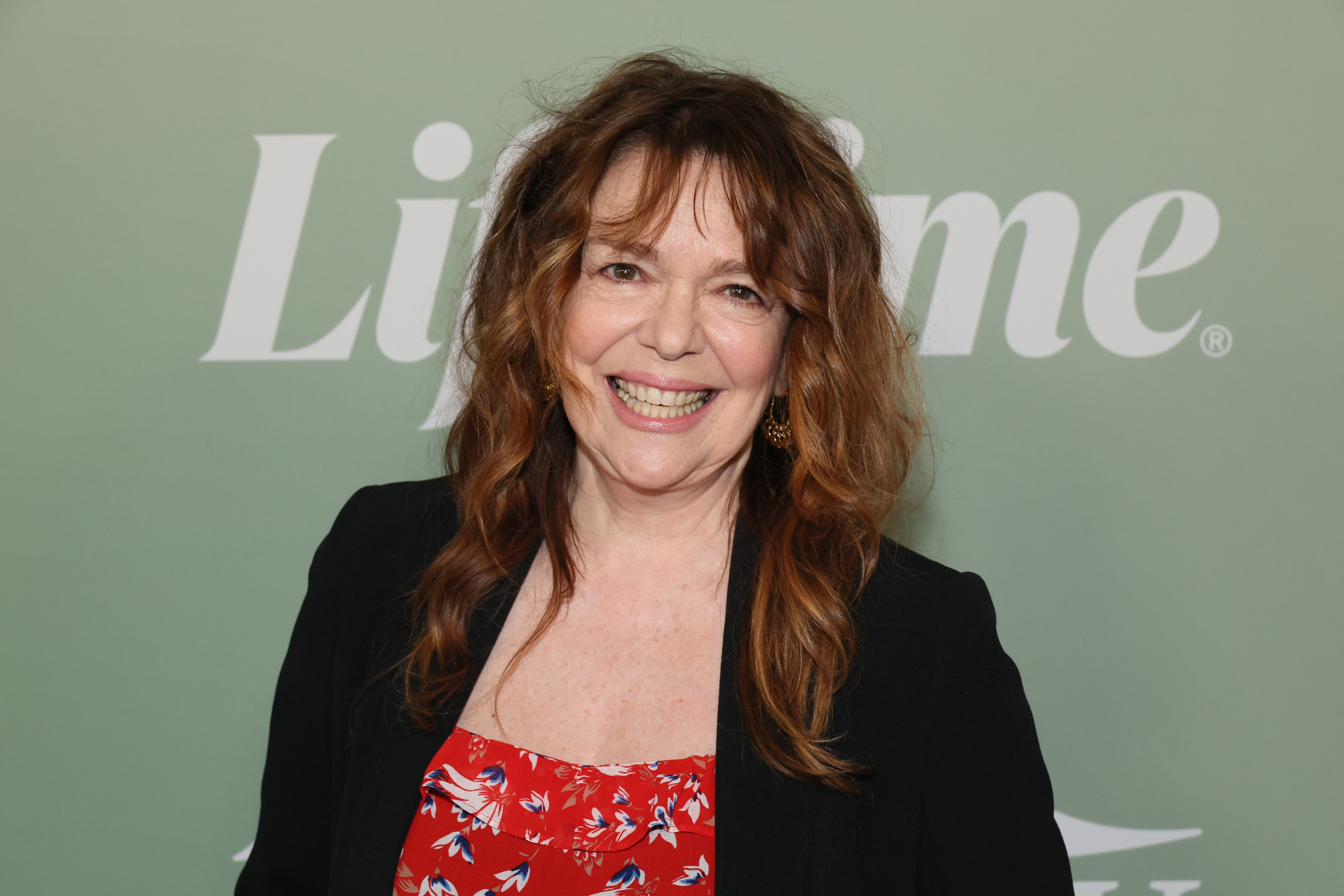Deirdre O'Connell poses in front of a Lifetime backdrop, wearing a floral print dress and a black blazer. She is smiling broadly