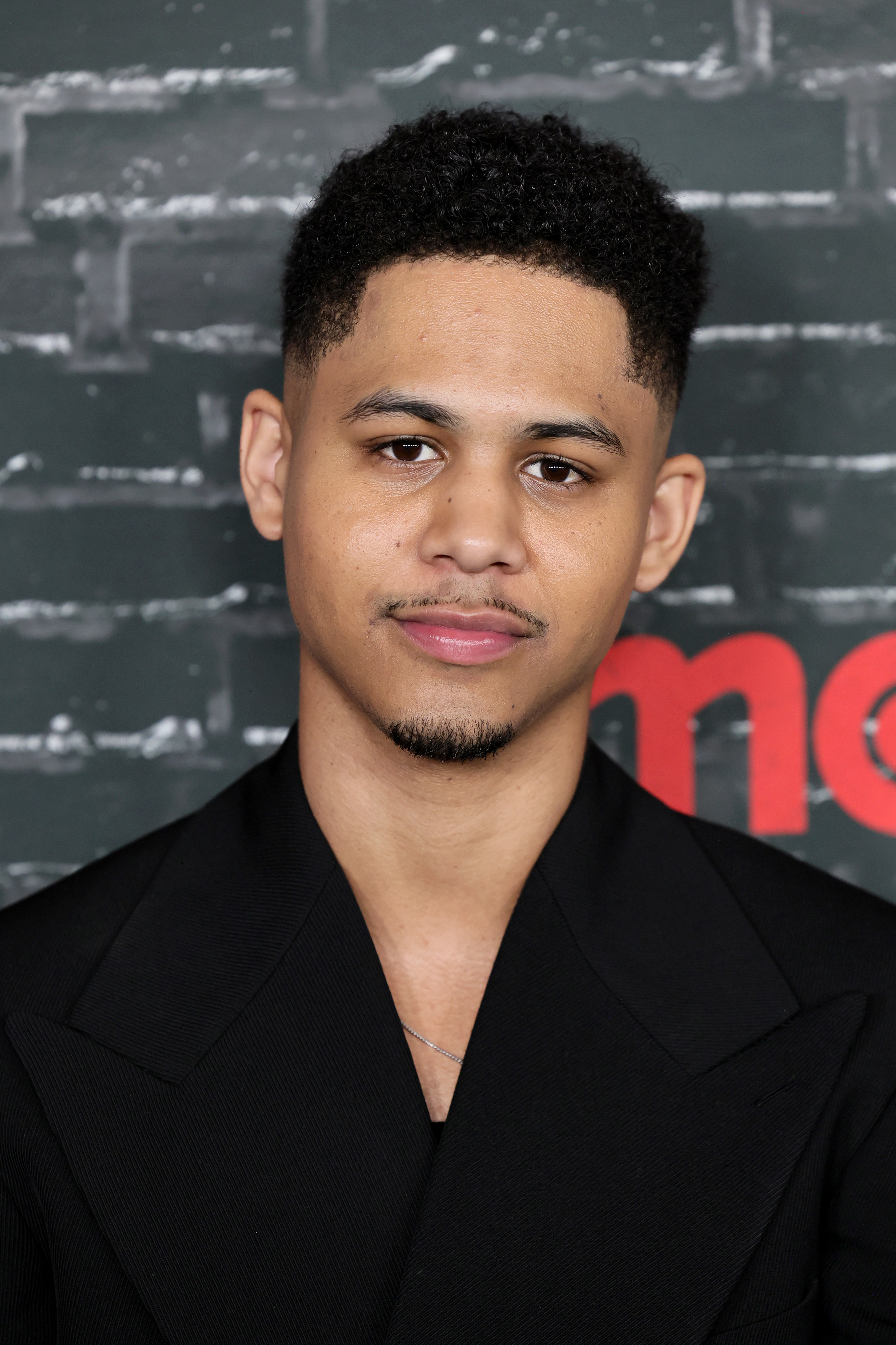 Rhenzy Feliz dressed in a black jacket stands in front of a dark brick wall backdrop, looking at the camera