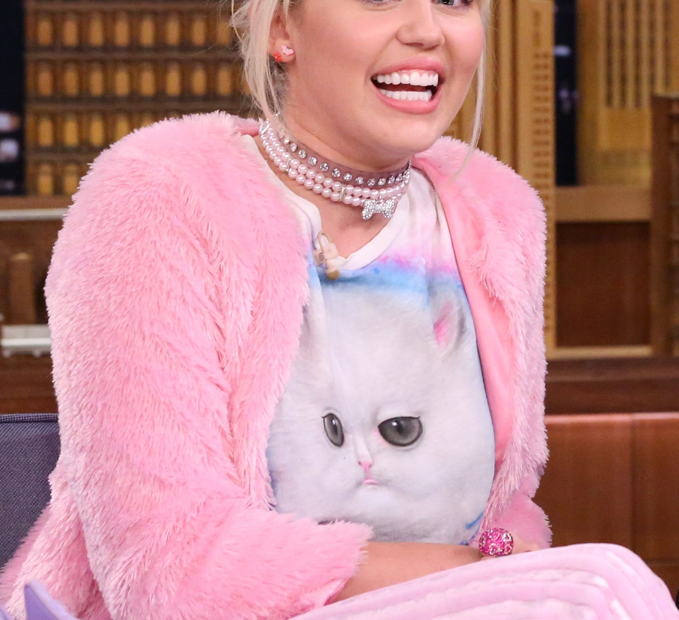Miley Cyrus is seated on a talk show set wearing a fluffy outfit, featuring a cat-themed shirt, with cityscape props in the background. Her hair is bleached blonde with dark roots, pulled back with bangs