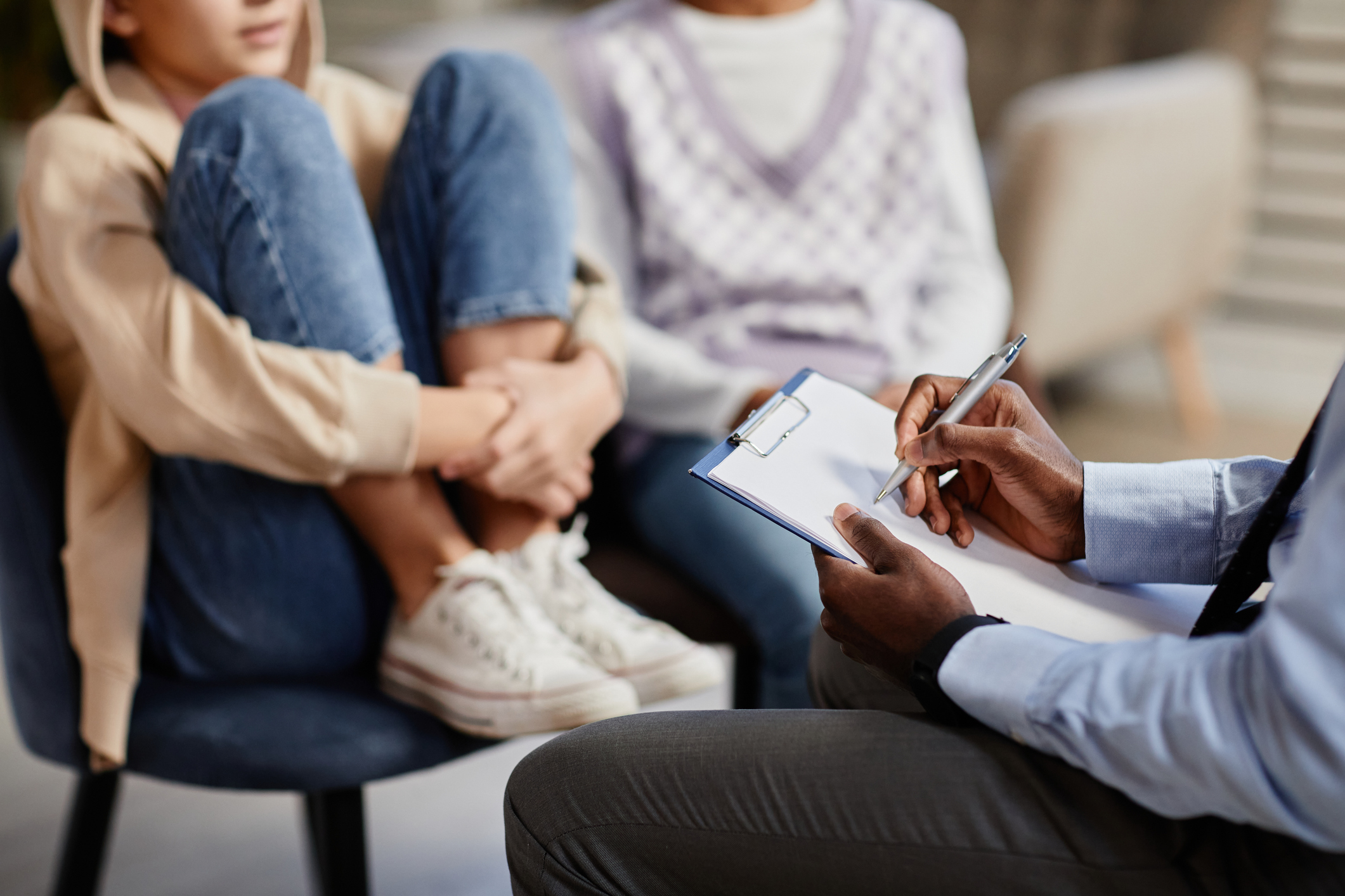 Two children sit on chairs while an adult holds a clipboard and takes notes, suggesting a consultation or counseling session