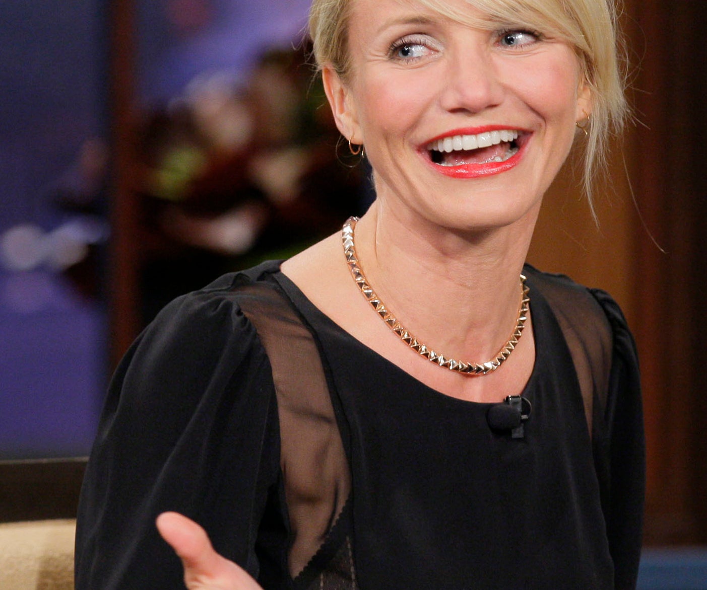 Cameron Diaz smiling while seated and gesturing during an interview. She is wearing a stylish dark dress and a gold necklace, and she has short hair pulled back