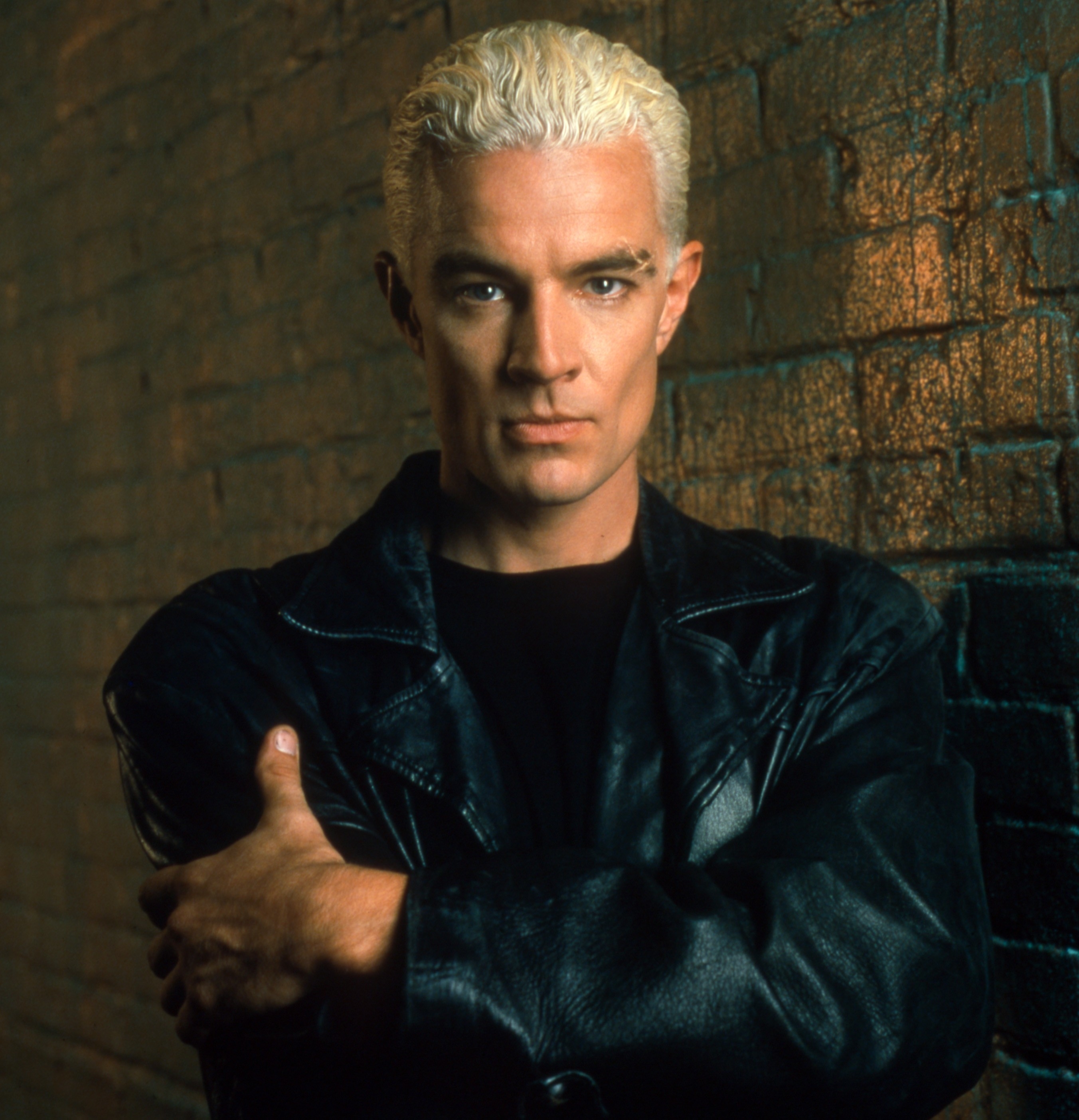 James Marsters poses against a textured brick wall, wearing a leather jacket and a stern expression. His bleach blonde hair is short and styled up
