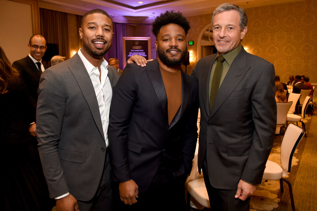 Michael B. Jordan and Ryan Coogler pose together at a formal event. Other attendees and tables are seen in the background