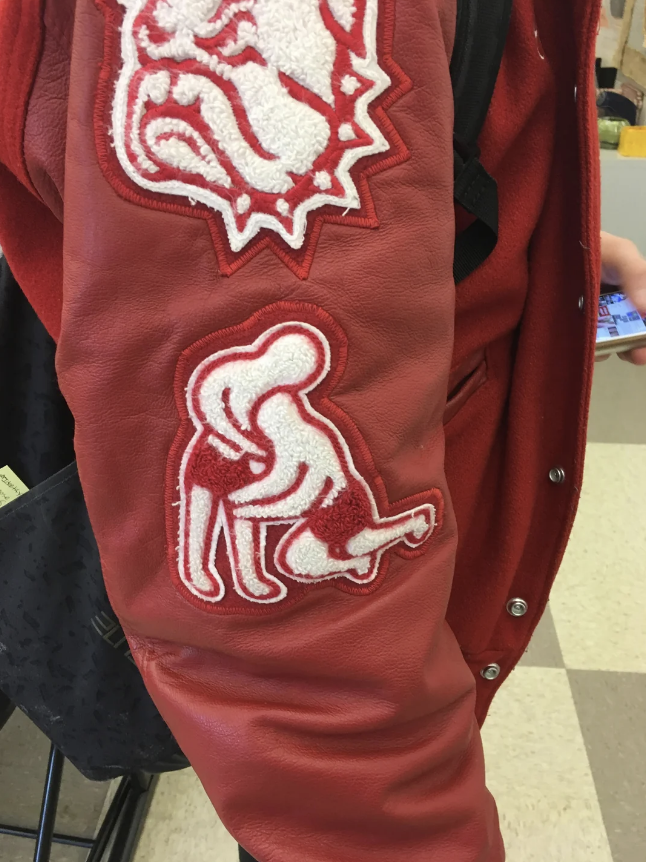 A person is wearing a red jacket with two white patches: one of a bulldog and another depicting a wrestling scene. The person is holding a smartphone