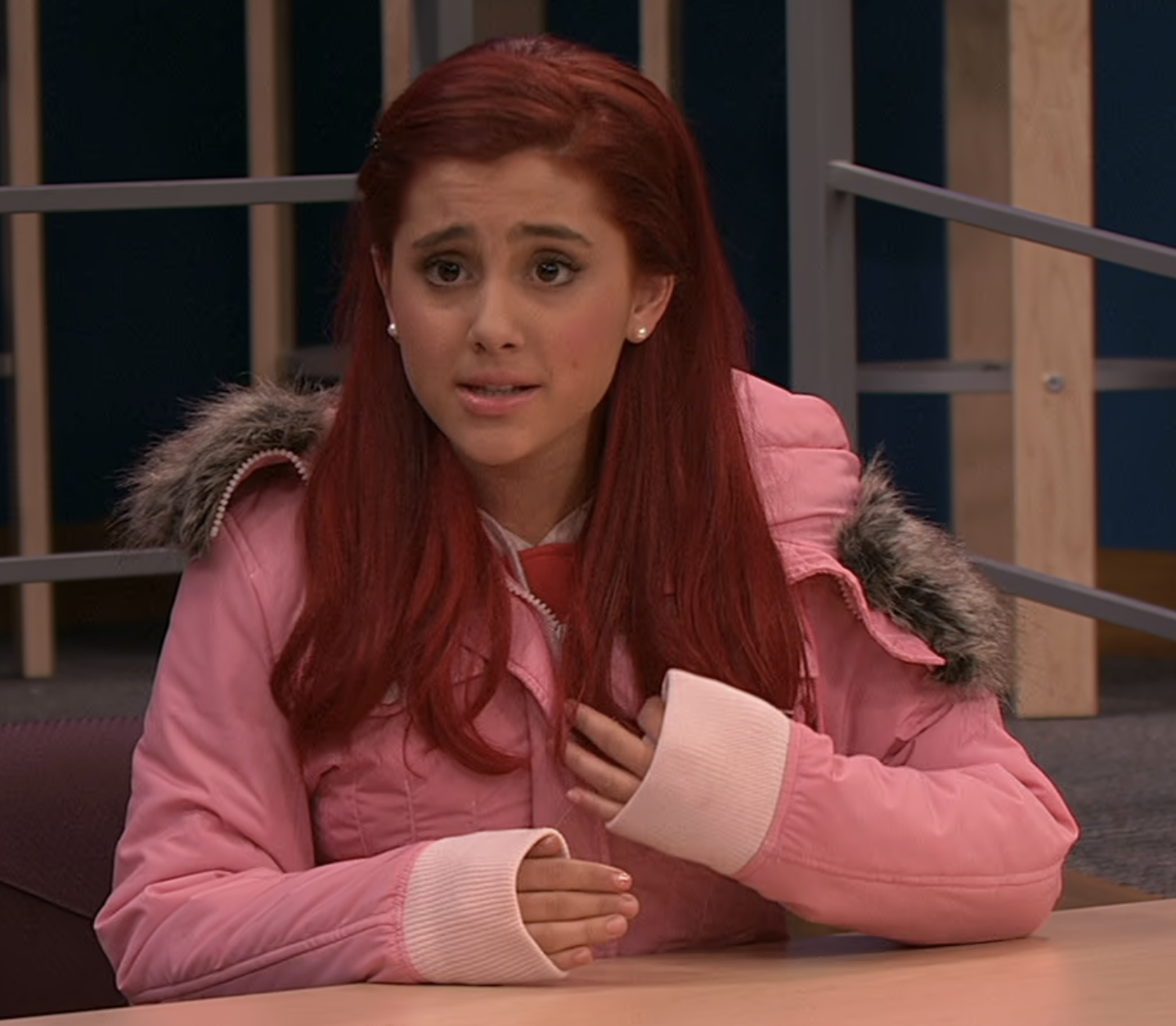 Ariana Grande sits at a table wearing a jacket with fur trim, with long hair dyed a bold, unnatural color