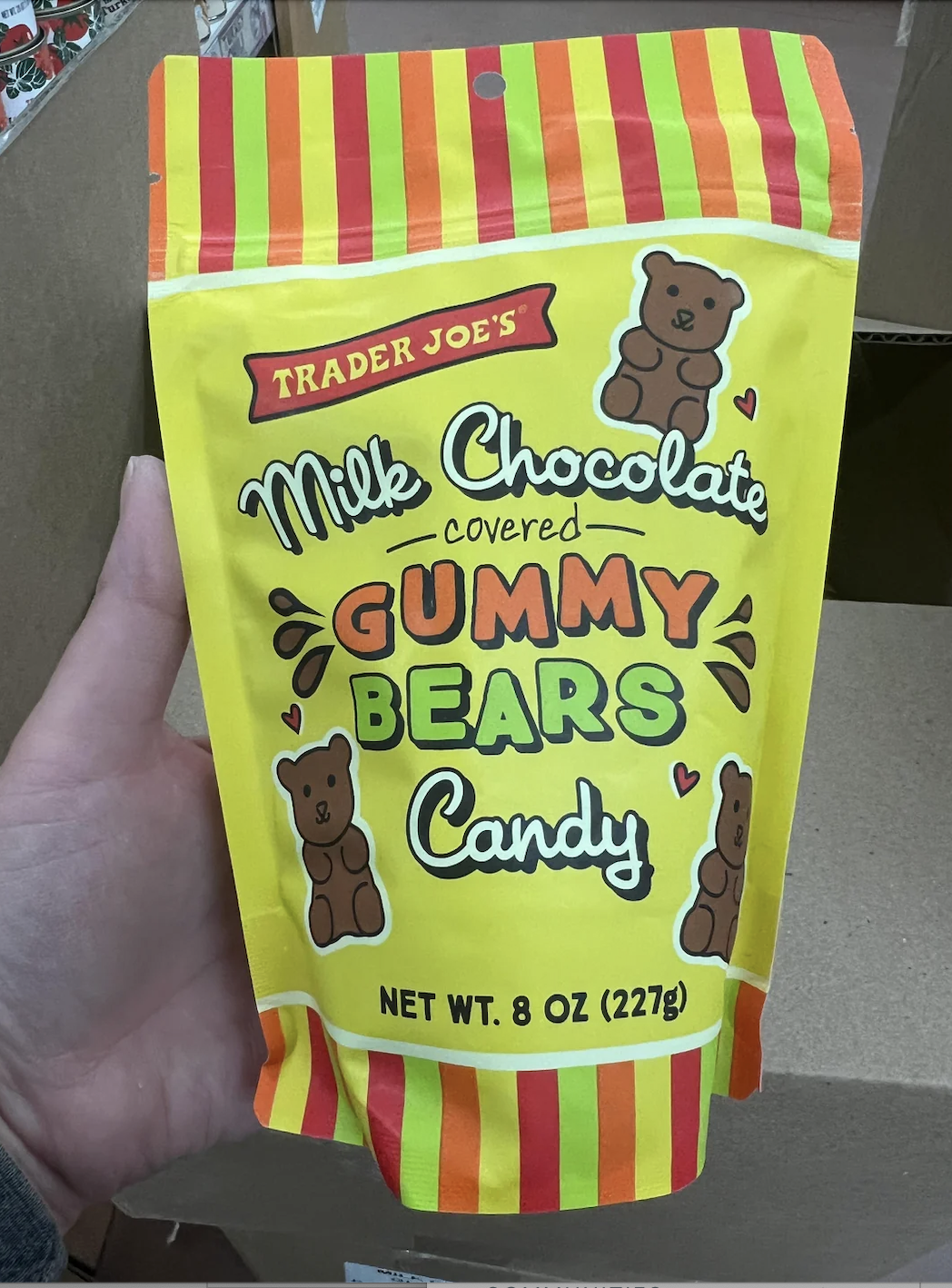 A person's hand holds a bag of Trader Joe's Milk Chocolate Covered Gummy Bears Candy, 8 oz (227g). The bag is decorated with bears and striped patterns