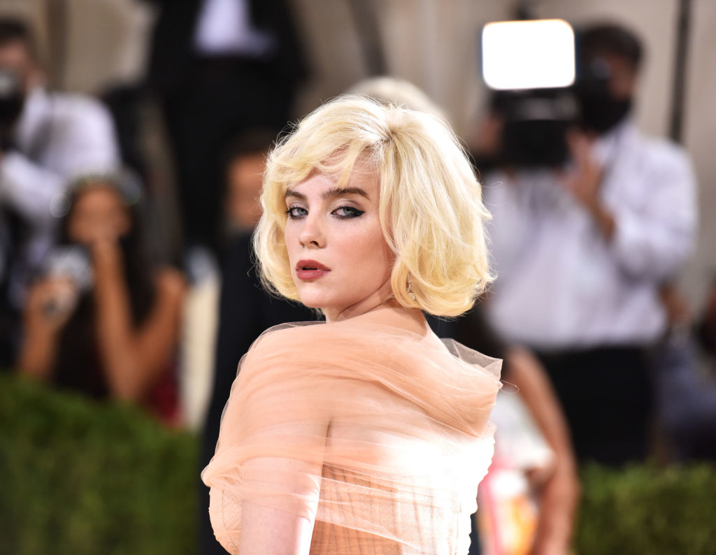 Billie Eilish on the red carpet, wearing an elegant, off-the-shoulder gown with wavy, layered hair and striking makeup