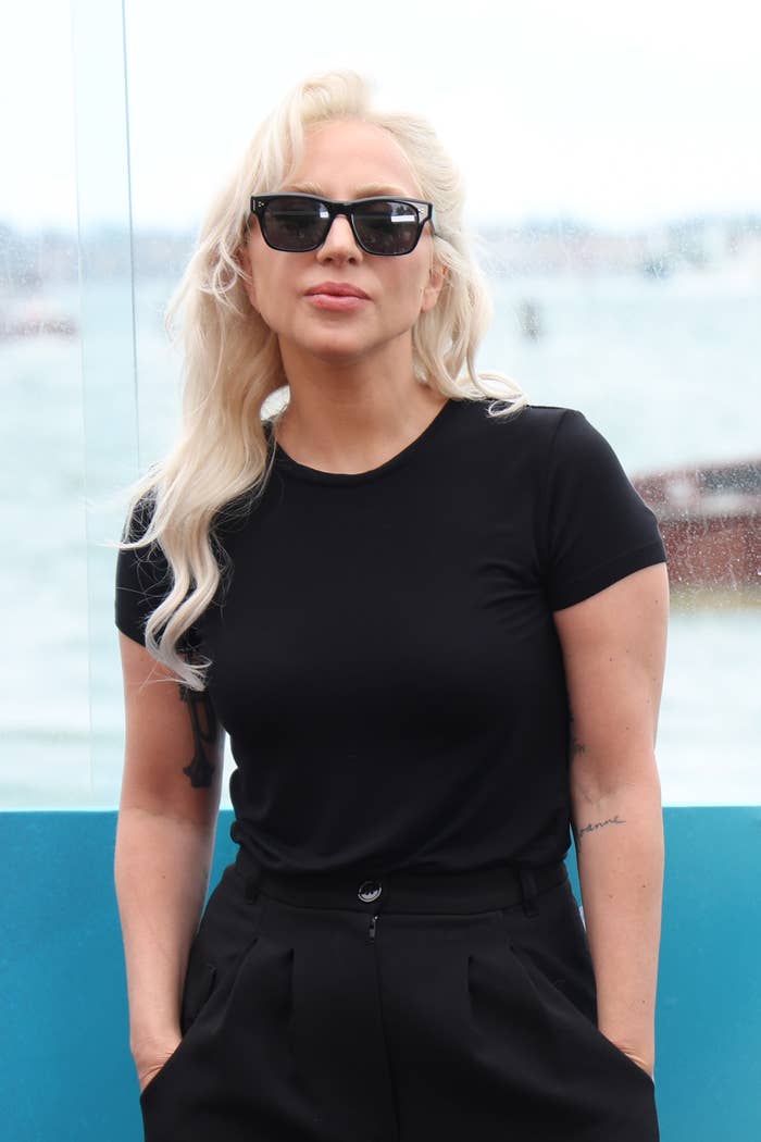 Lady Gaga is wearing sunglasses, a dark t-shirt, and high-waisted pants, standing in front of a waterfront