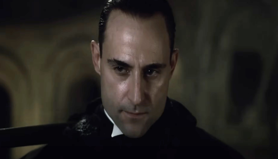 Actor Mark Strong in character, wearing a dark, formal attire with a serious expression in a scene from a movie