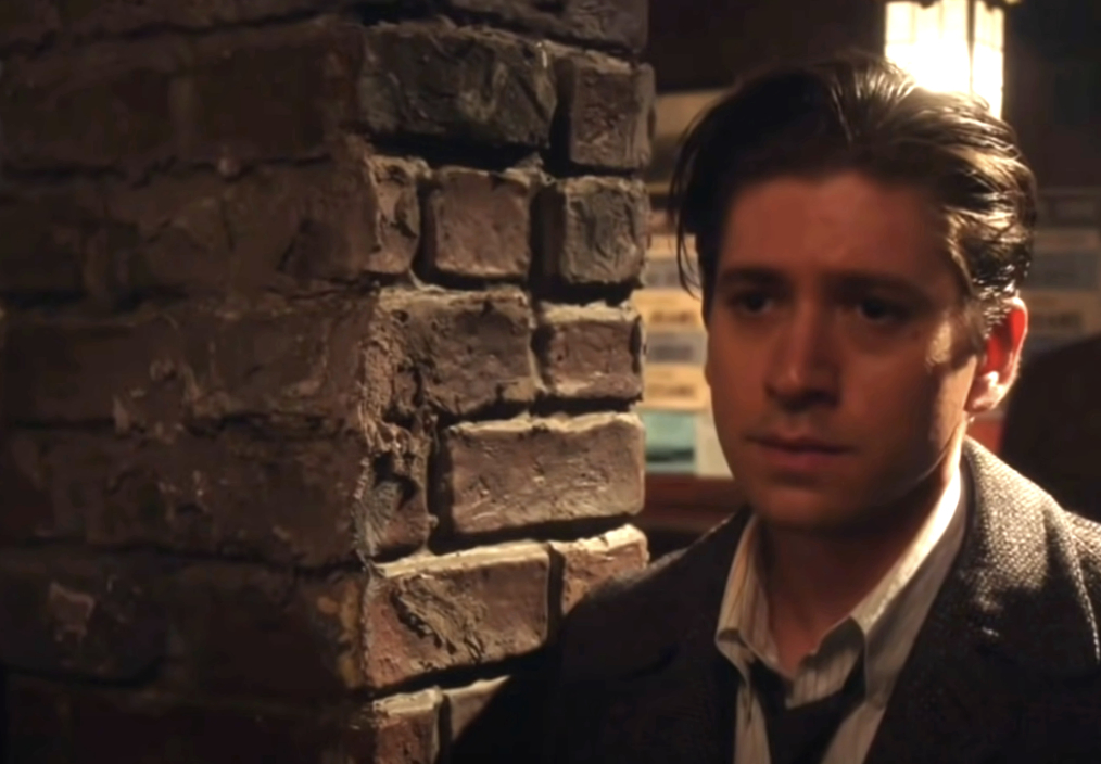Michael Zegen, in mid-20th-century attire, stands pensively by a brick wall in a likely TV or movie scene