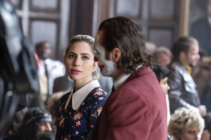 Lady Gaga, in a floral dress with a white collar, looks thoughtfully at Joaquin Phoenix, dressed in a maroon suit jacket, in a busy indoor setting in a scene from the sequel
