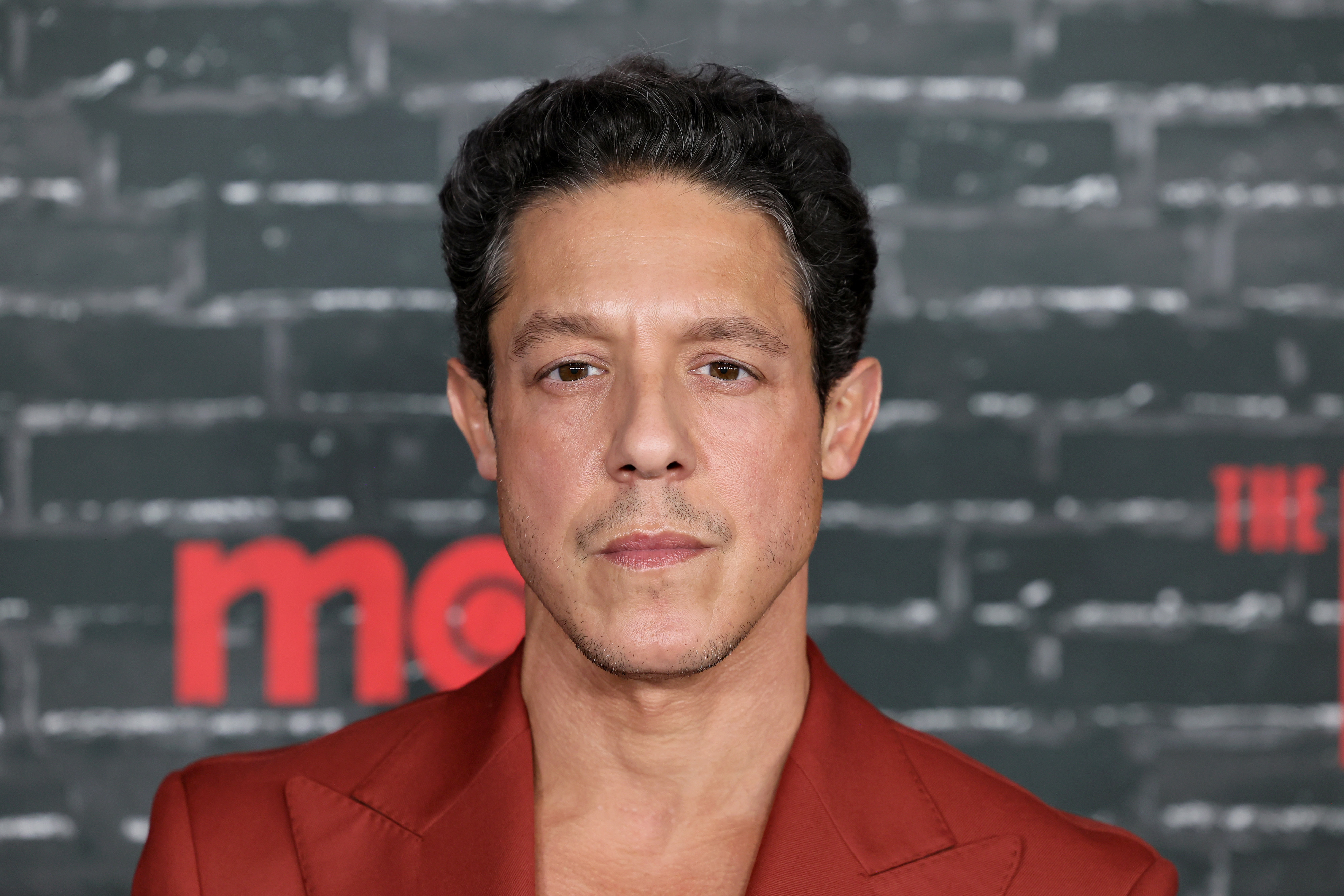 Benjamin Bratt attends an event. He wears a tailored suit jacket with a plain shirt underneath. The background has a brick wall pattern