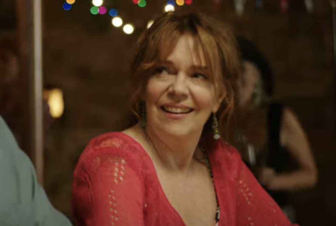 Woman with tied-back hair smiling, wearing a lace-style top, in a lively setting with blurred background lights